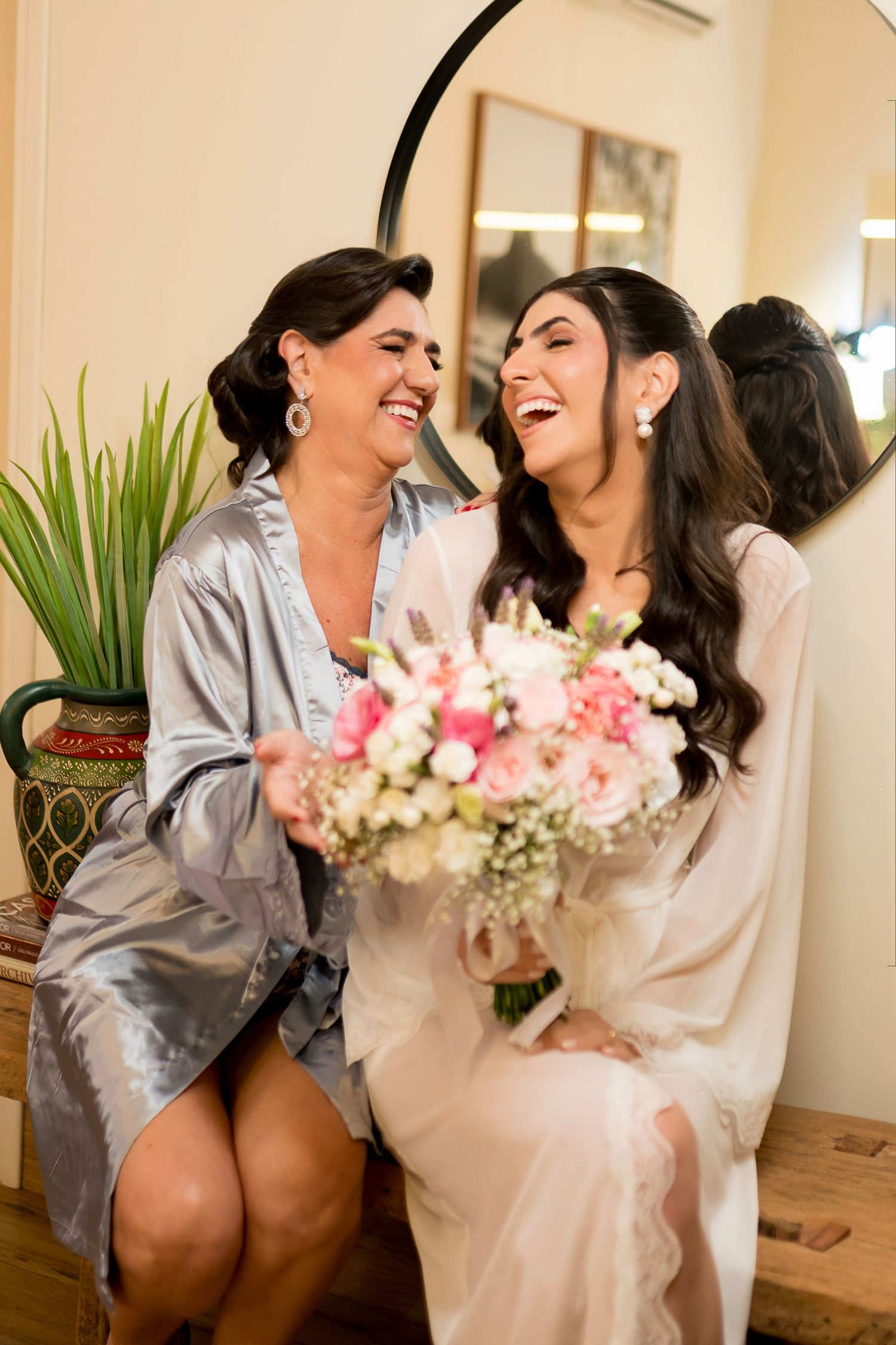 Bride in white robe laughing with bridesmaid in silver robe while holding pink and white bouquet.