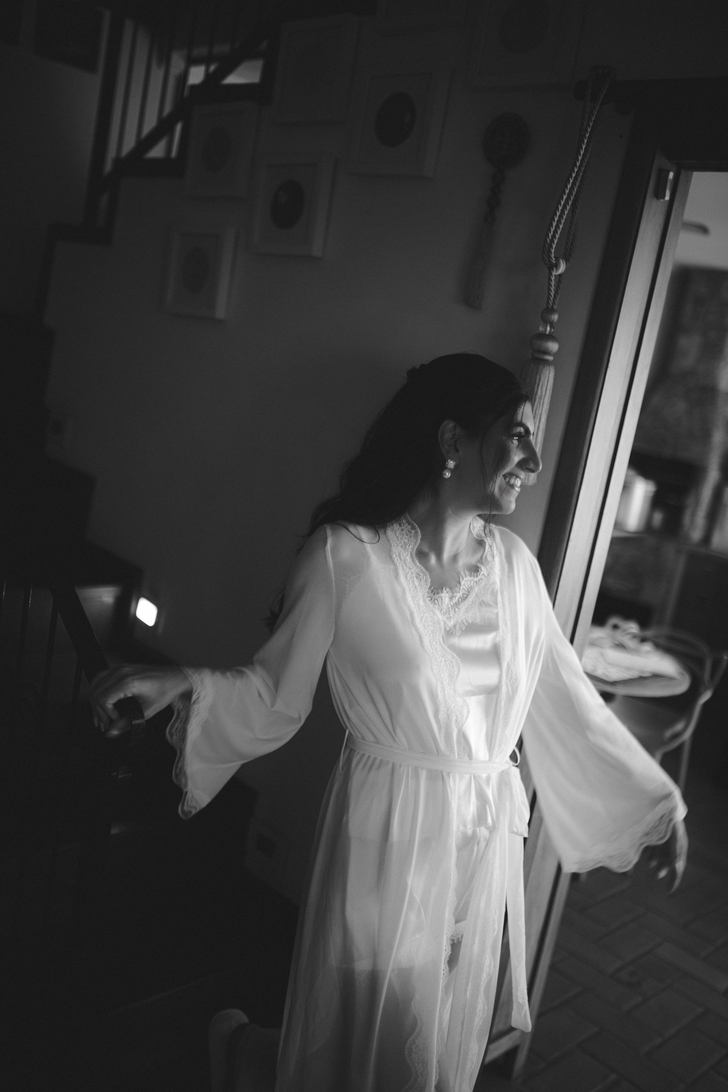 Black and white photo of a smiling bride in a sheer white robe standing by a doorway in soft natural light.