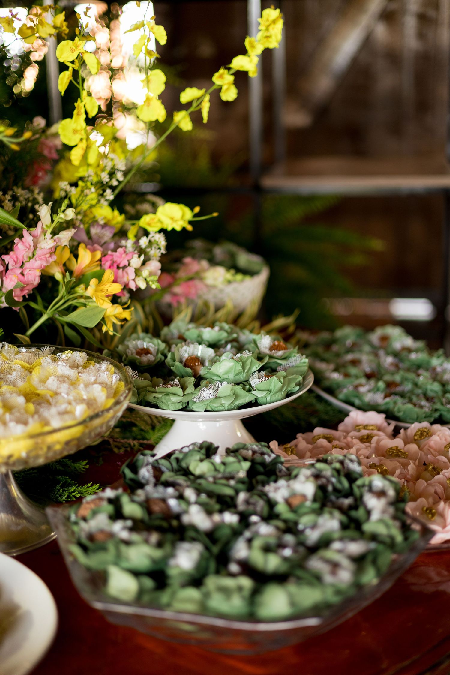 Elegant dessert table with green and pink sweets on tiered stands, surrounded by colorful floral arrangements.