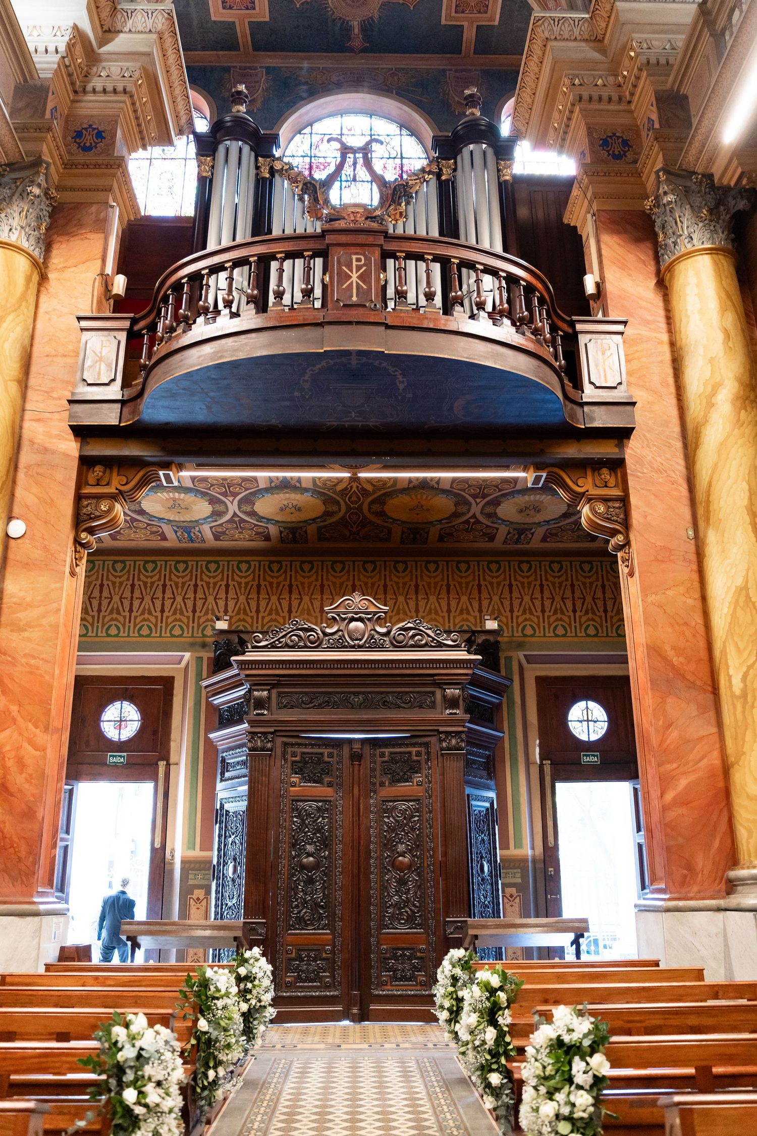 Ornate church interior with pipe organ, carved wooden doors, marble columns, and floral arrangements lining the aisle.