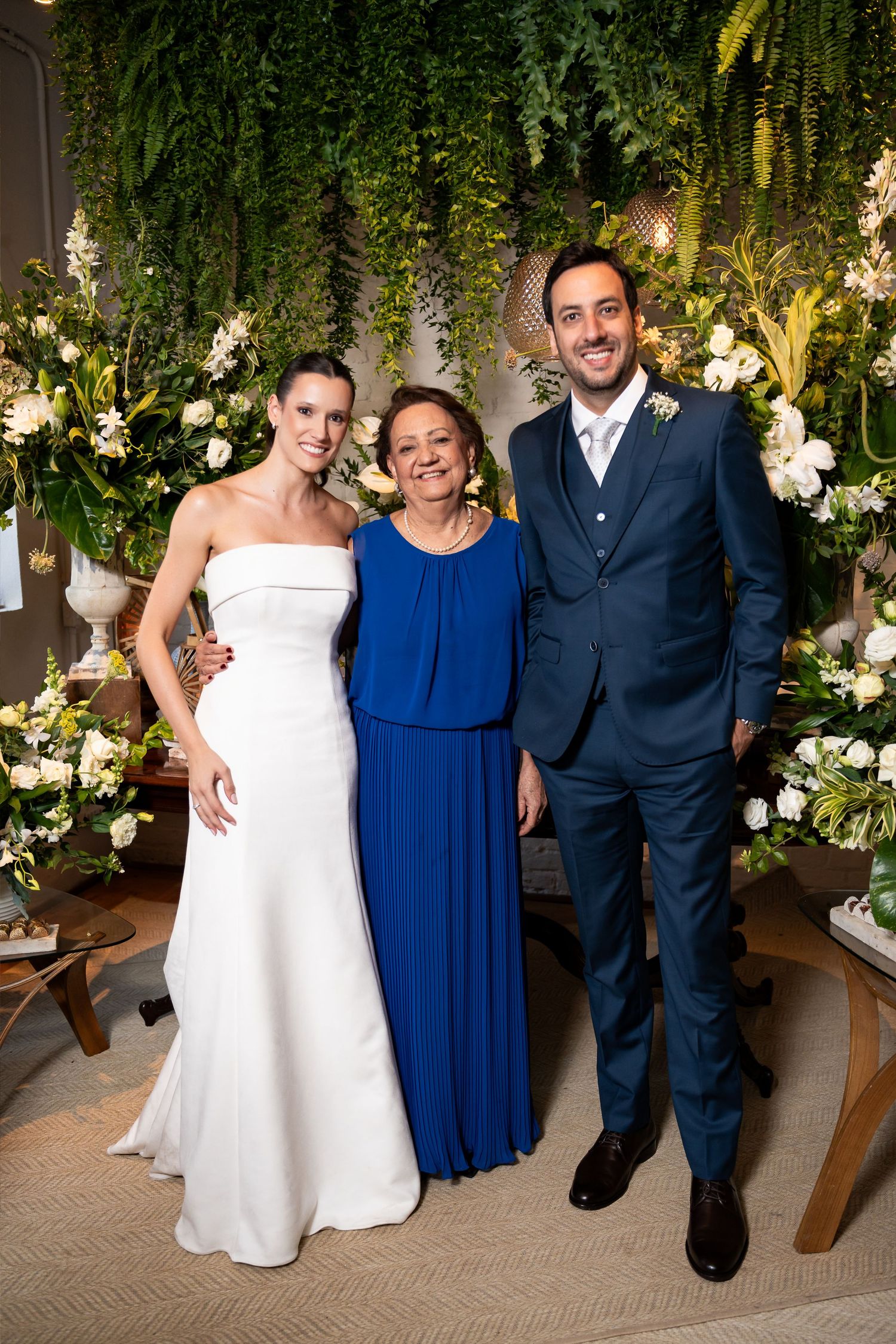 Bride in white gown, woman in blue dress, and groom in navy suit posing together at a floral wedding reception.