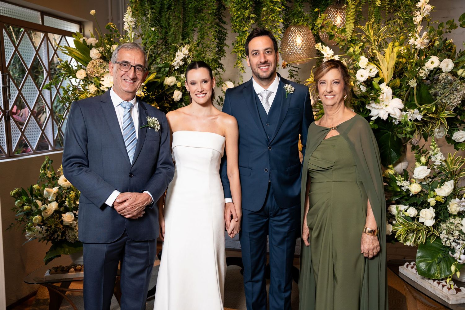 Wedding party of four posing together, bride in white gown, groom in navy suit, flanked by family near floral backdrop.