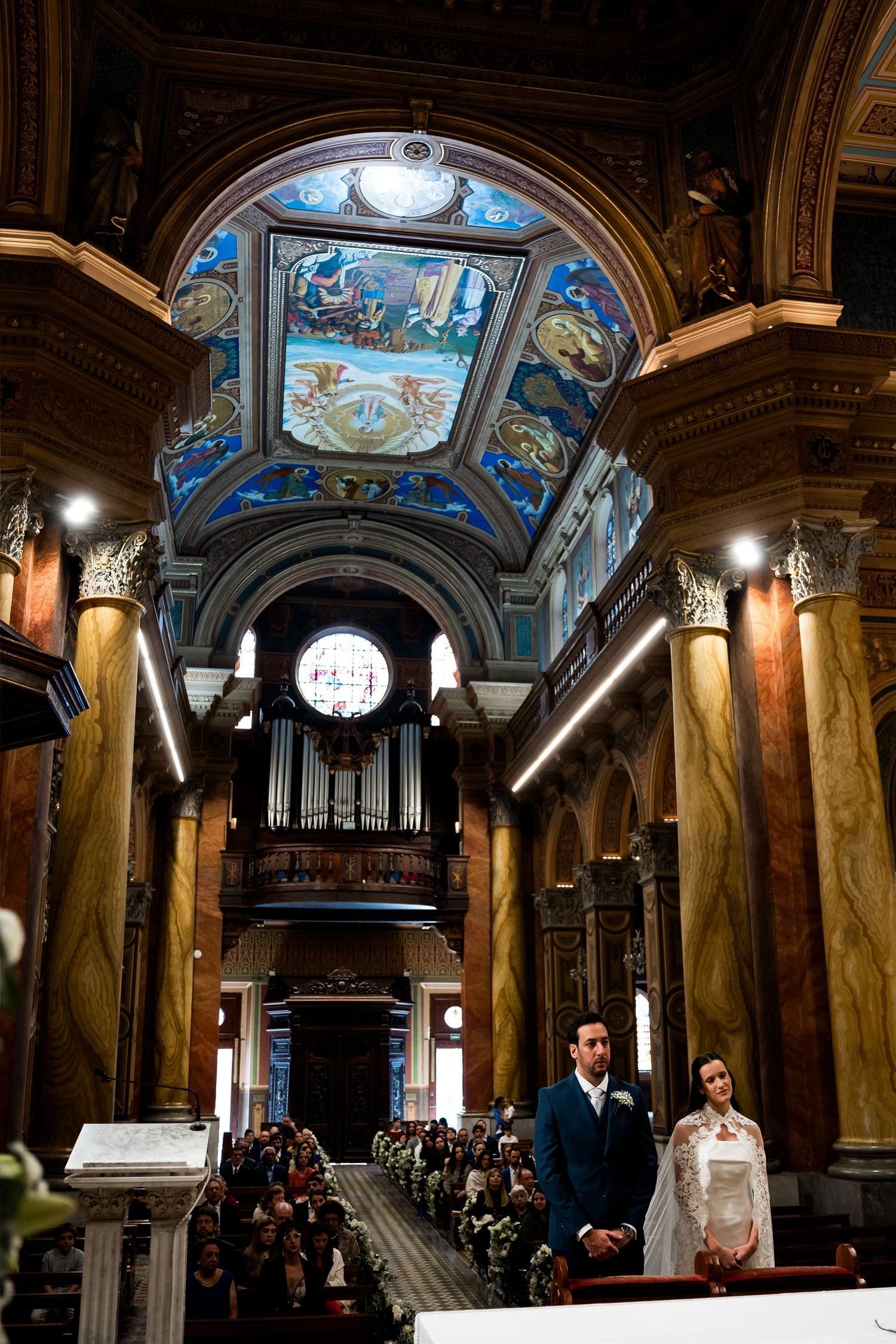 Couple stands at the altar of an ornate church with stunning blue frescoed ceilings and marble columns during wedding.