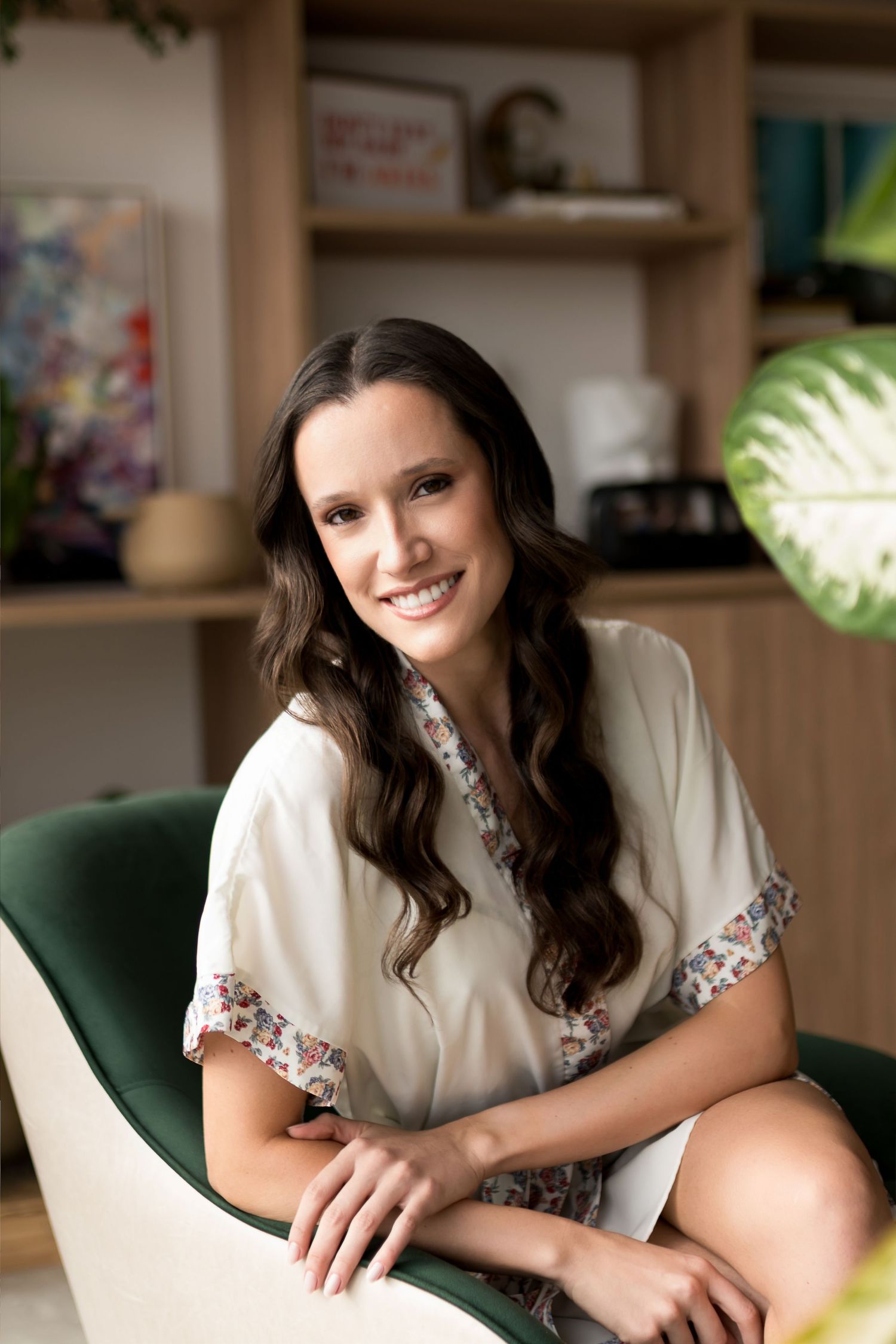 Smiling young woman with long dark wavy hair sitting in a green chair wearing a floral-trimmed white blouse indoors.