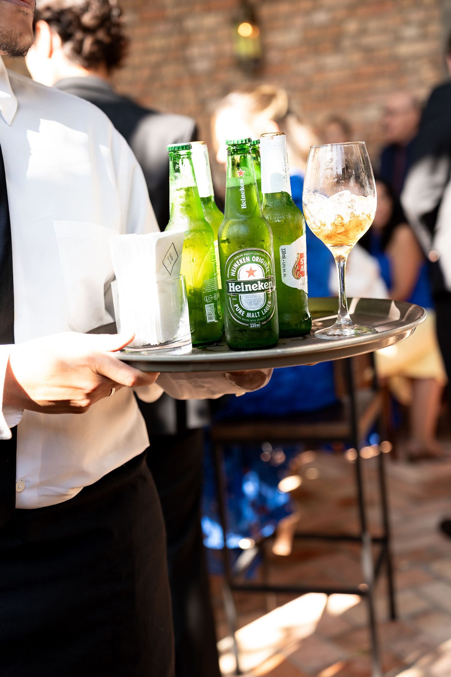 Waiter holding silver tray with Heineken beer bottles and wine glass at outdoor wedding reception.