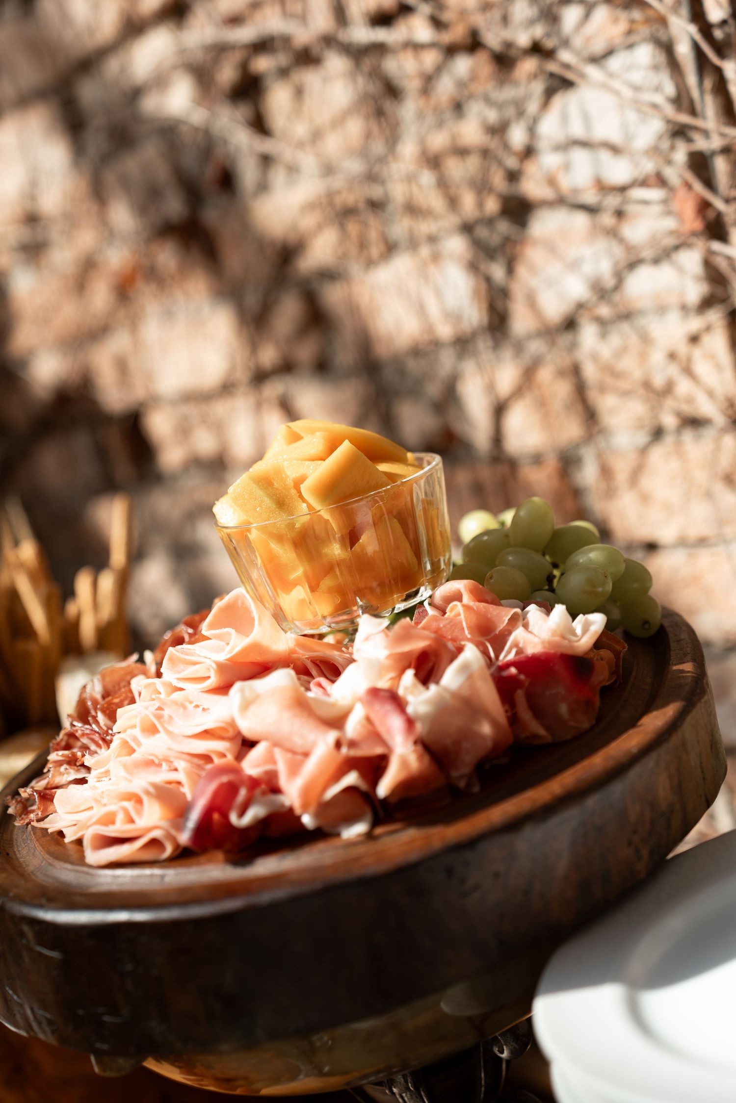 Rustic wooden platter with prosciutto, salami, green grapes, and a glass of melon at an outdoor restaurant.