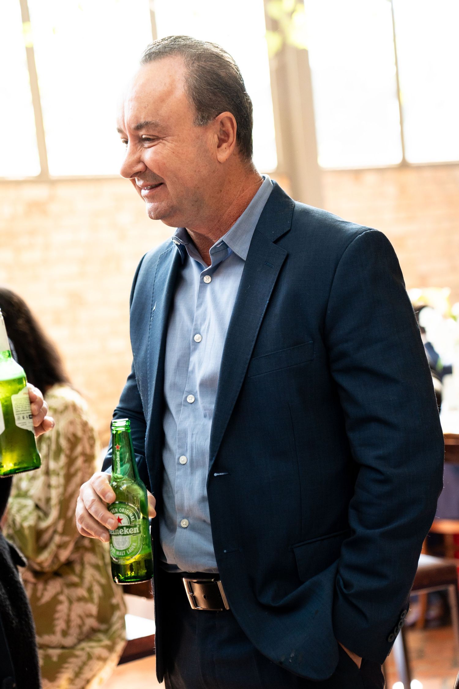 Smiling middle-aged man in navy blazer holding green beer bottle at social gathering.