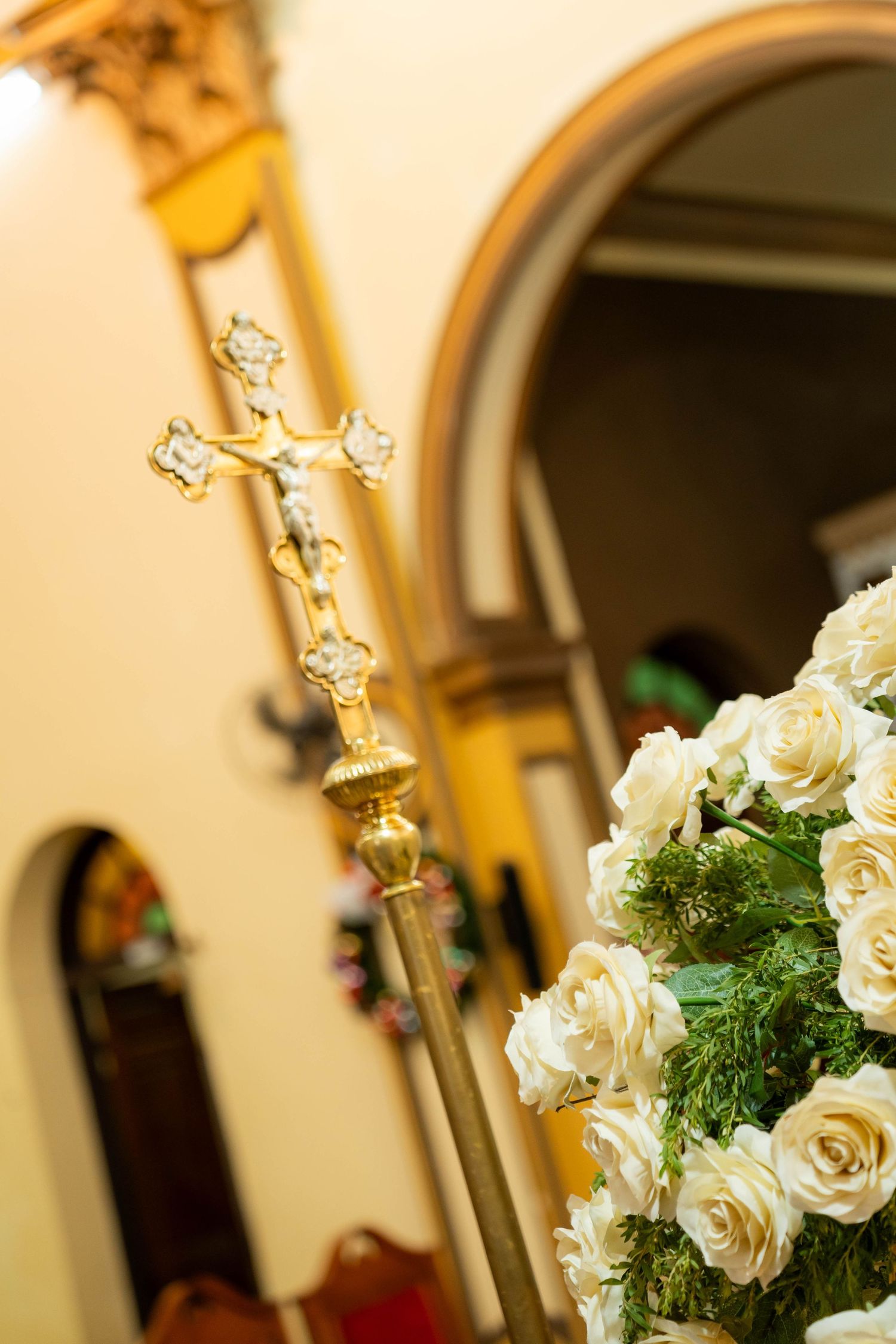 Golden crucifix on a processional staff beside white roses in a decorated church interior.