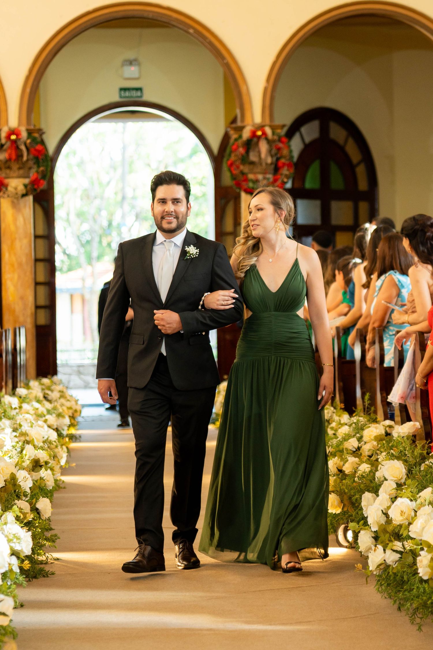 Smiling groomsman in black suit escorts bridesmaid in emerald green dress down floral church aisle.