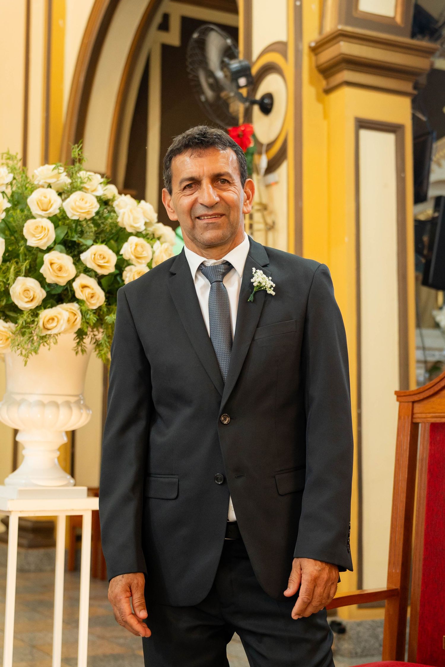 Smiling man in black suit with boutonniere stands near white roses at a wedding ceremony.