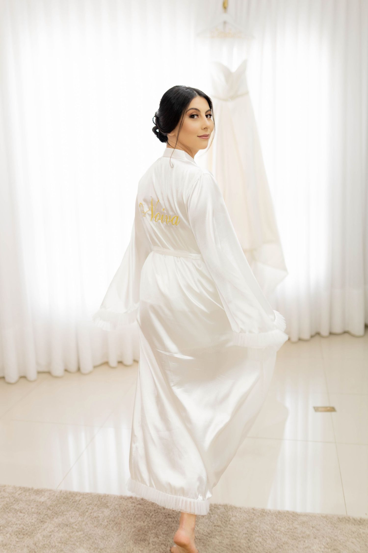 Bride in white satin robe looking over shoulder near wedding dress hanging by sheer curtains.