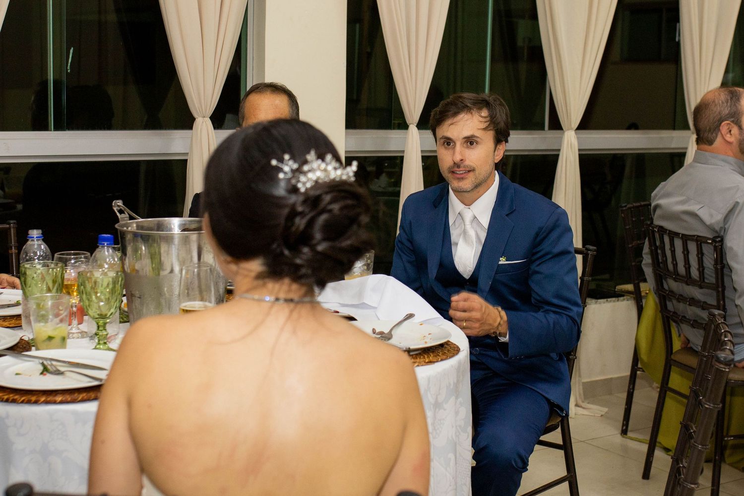 A groom in a blue suit sits at a wedding reception table, engaged in conversation with the bride in a white dress.