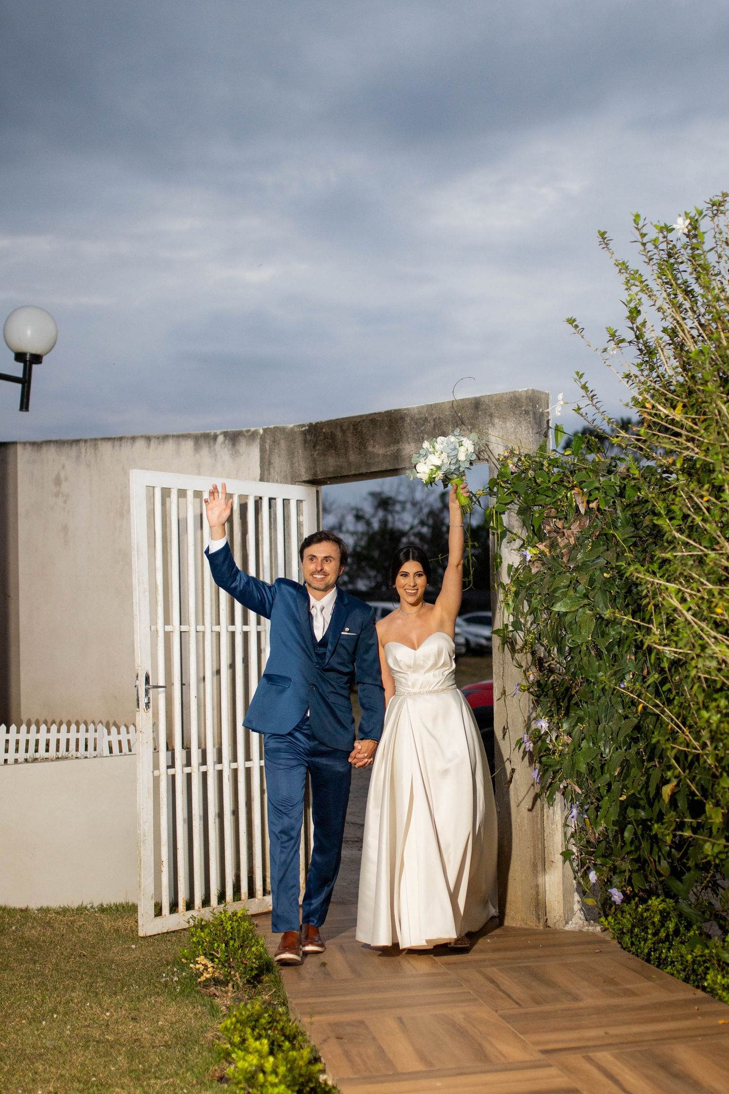 Bride and groom celebrating their wedding, raising hands joyfully, bride holding bouquet, groom in blue suit, outdoor venue.