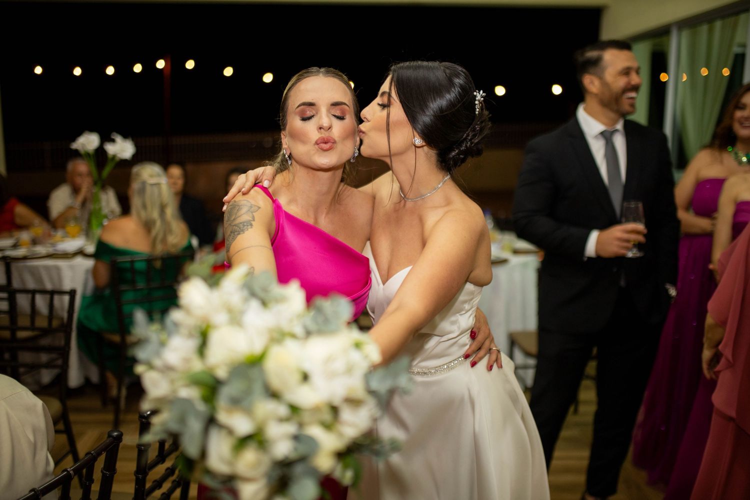 Bride in white gown shares a kiss on the cheek with a guest in a pink dress at an elegant wedding reception.