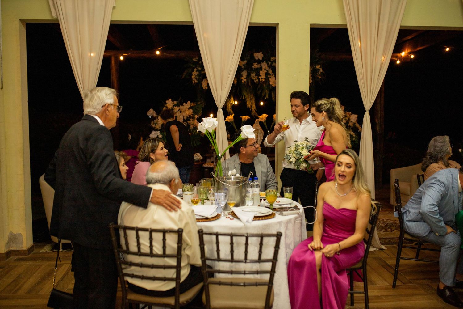 Elegant wedding reception with guests in formal attire seated at round tables adorned with floral centerpieces.