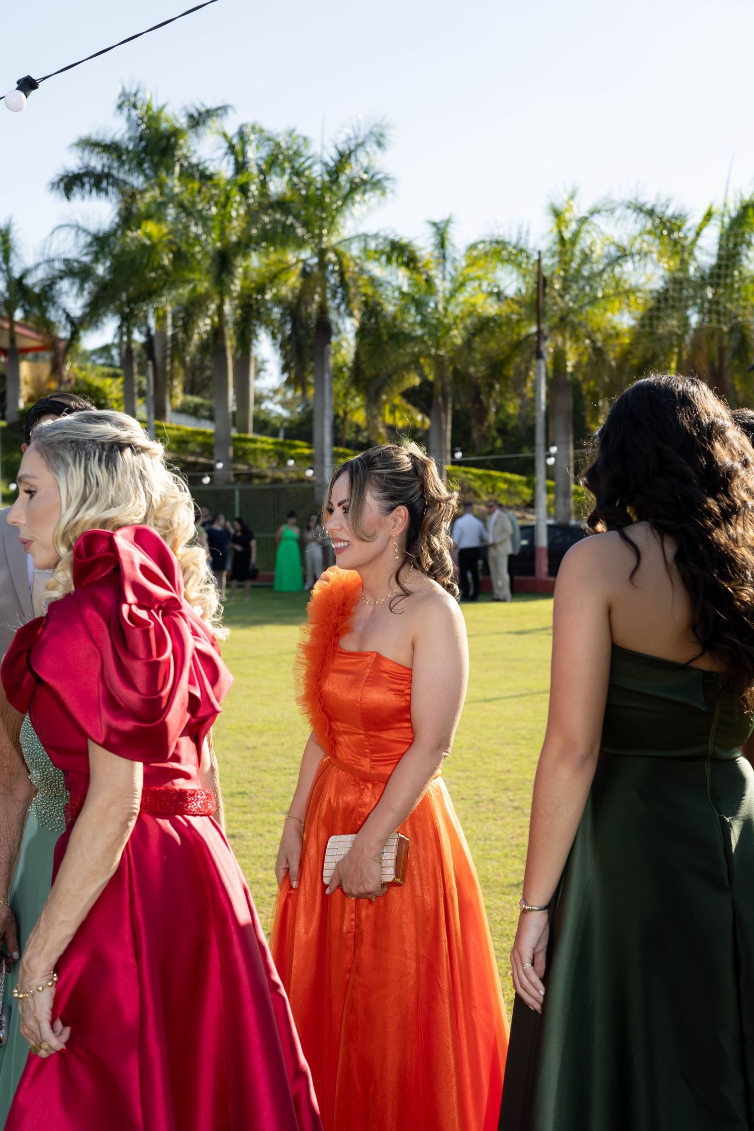 Women in elegant red, orange, and green formal gowns mingle on lawn at outdoor prom or gala event.