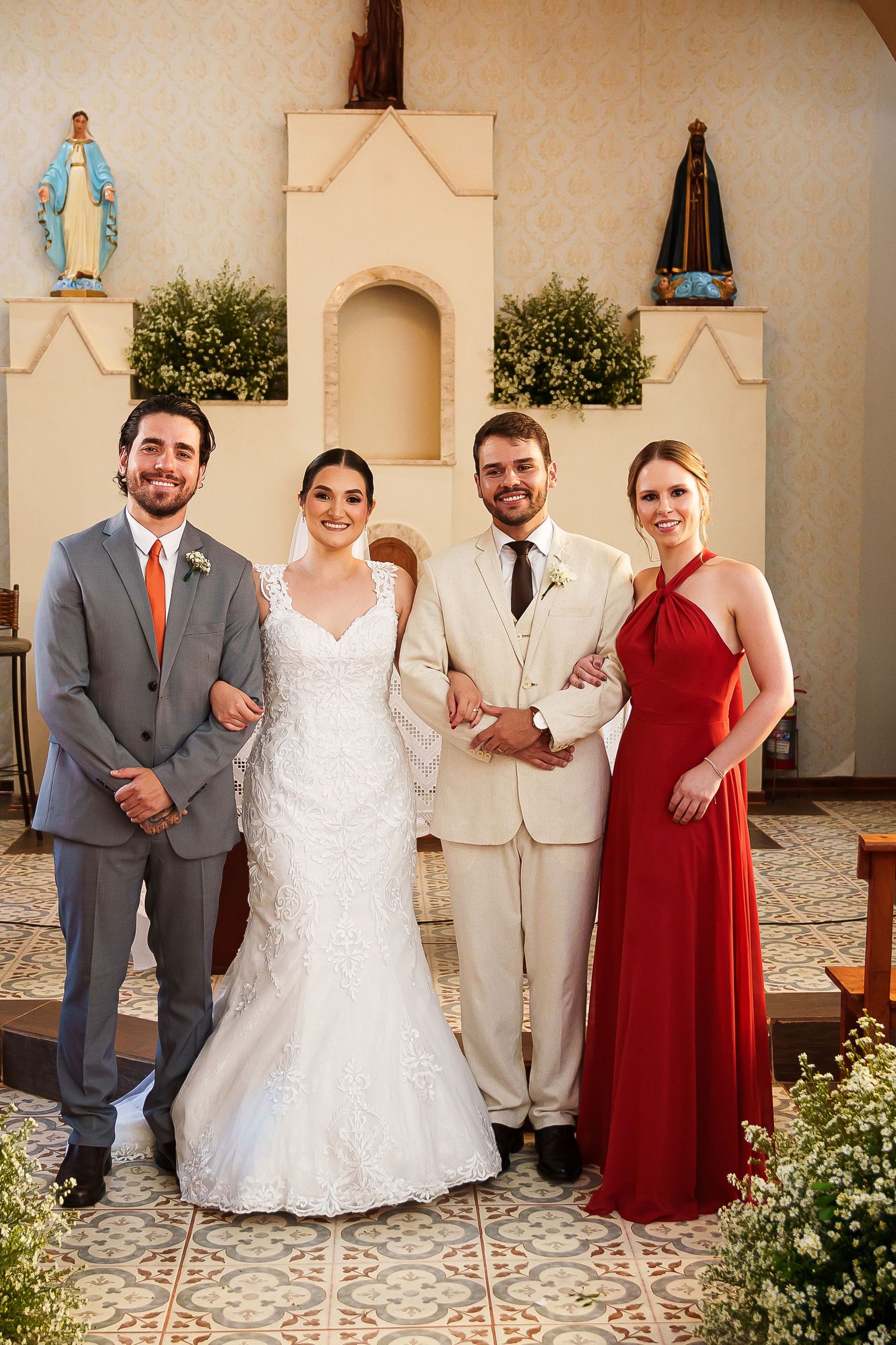 Wedding party posing in church, bride in white gown, groom in cream suit, groomsman in gray, bridesmaid in red dress.