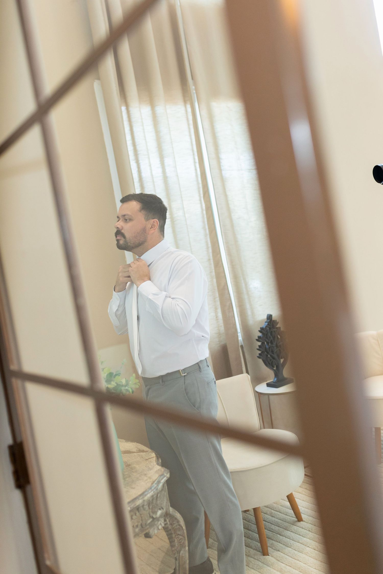 Man in white shirt adjusting his tie in a bright, elegant room, viewed through a glass-paned door.