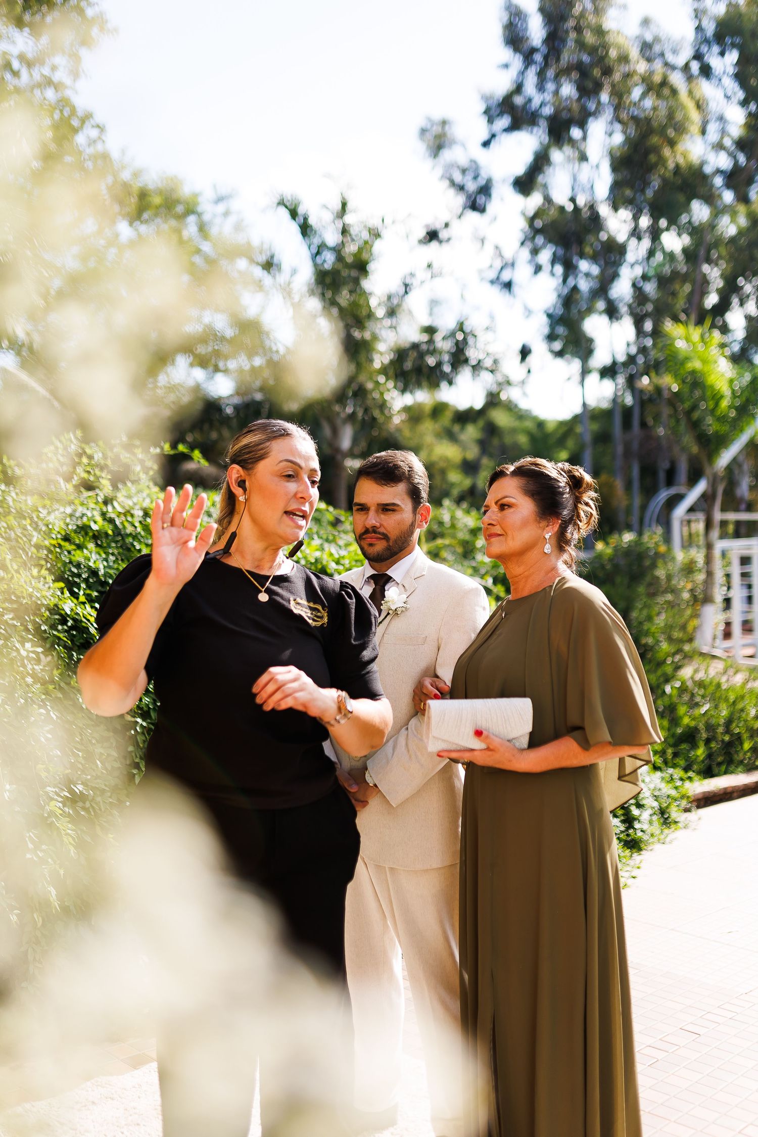 Wedding officiant in black gestures while groom in tan suit and bridesmaid in olive dress listen outdoors.