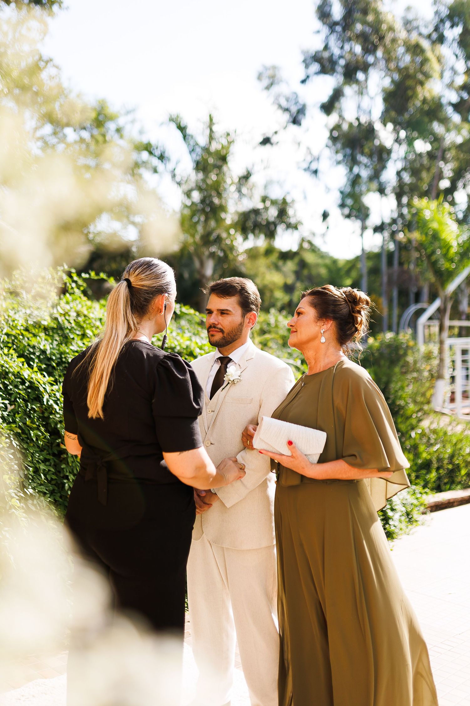 A groom in a white suit stands with two women in a sunlit garden during an outdoor wedding ceremony.