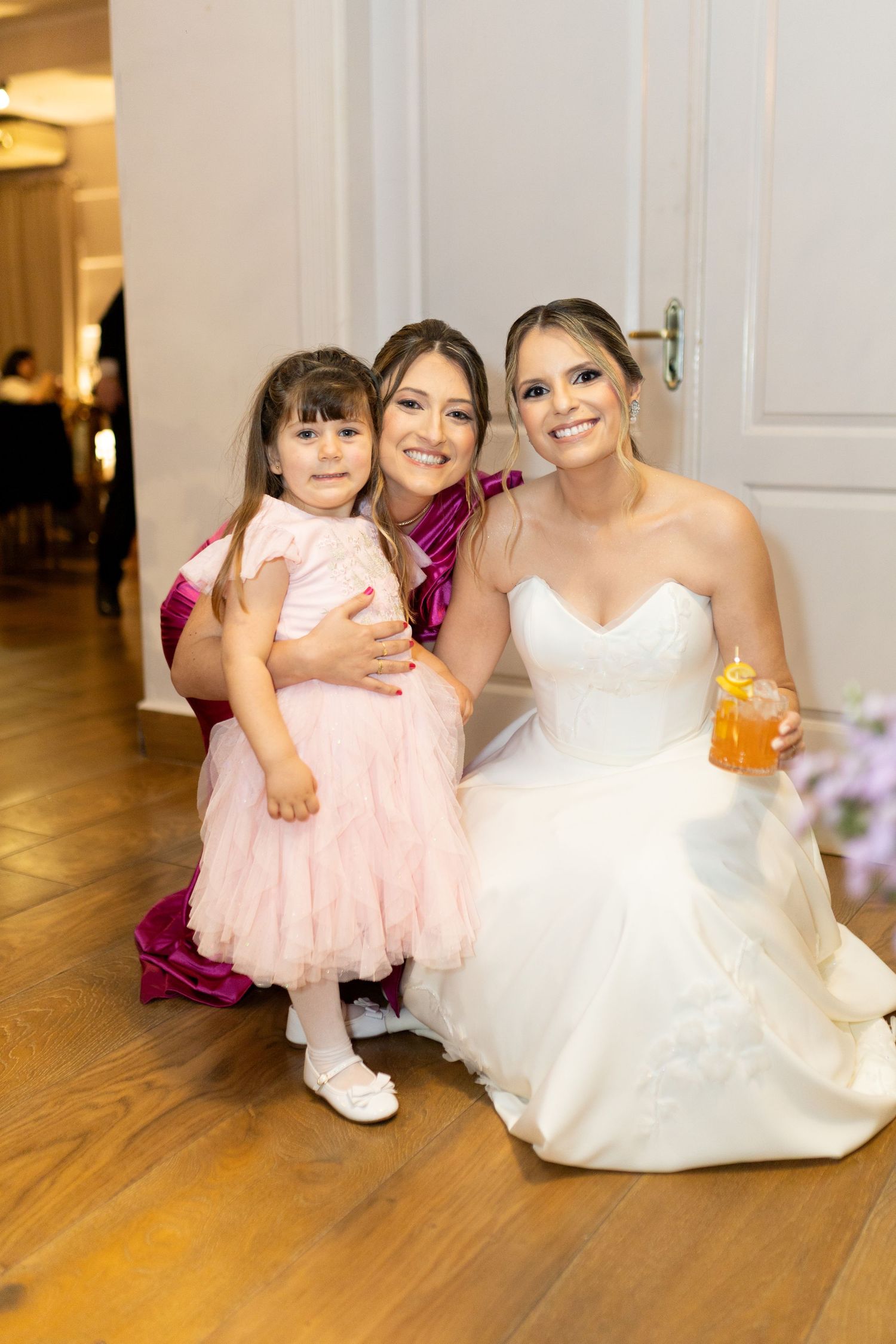 A bride in a white gown poses with a woman in magenta and a young girl in pink at a wedding reception.