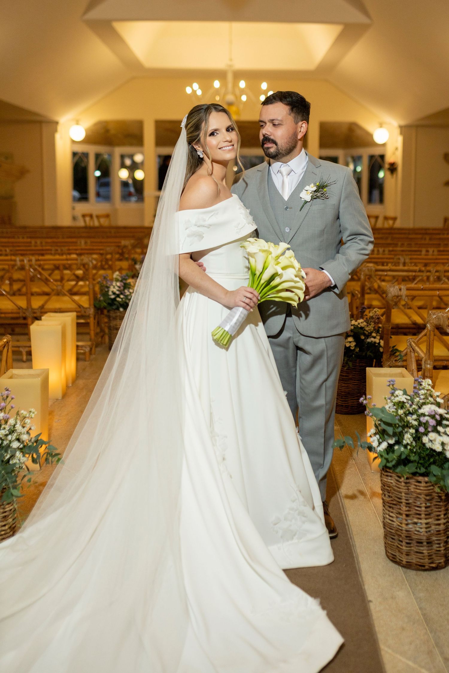 Bride in white off-shoulder gown with veil holds bouquet beside groom in gray suit in elegant chapel aisle.