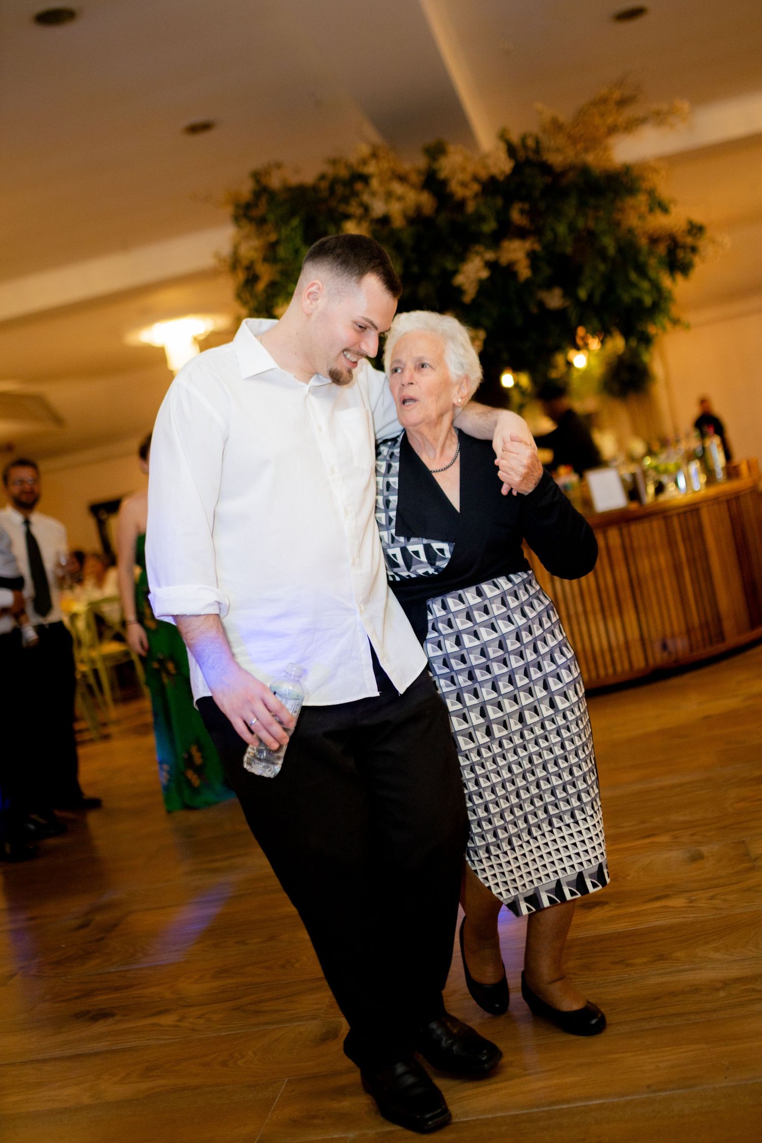 A young man in a white shirt dances with an elderly woman in a patterned dress at a wedding reception.