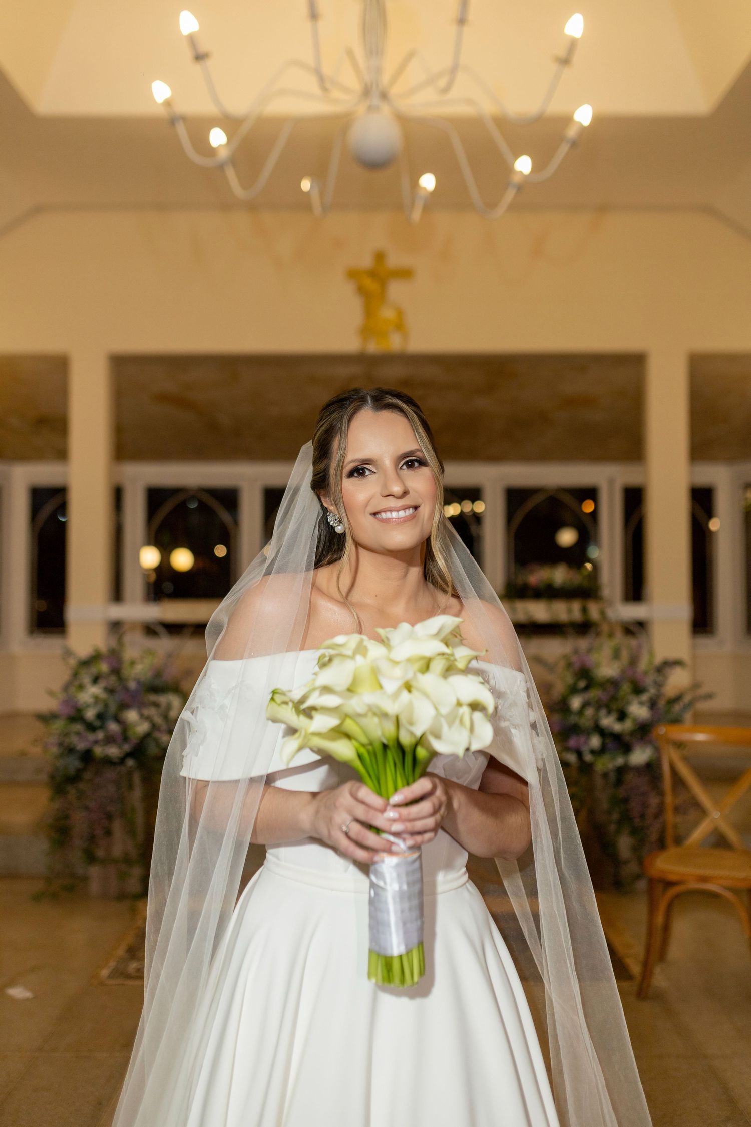 Smiling bride in white off-shoulder gown and veil holds calla lily bouquet inside elegant church with chandelier.