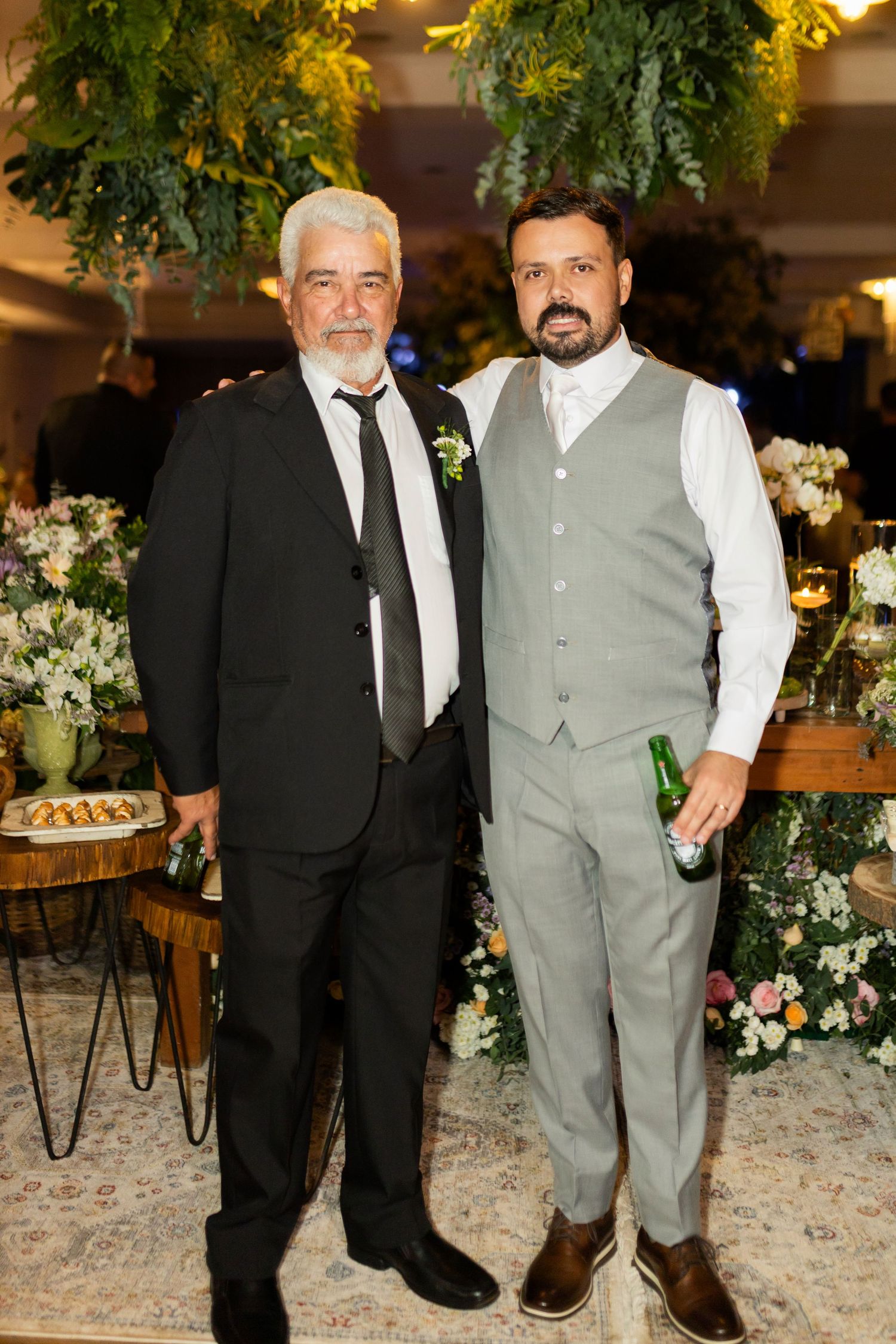 Two men in formal attire pose together at an elegant wedding reception with floral decorations.