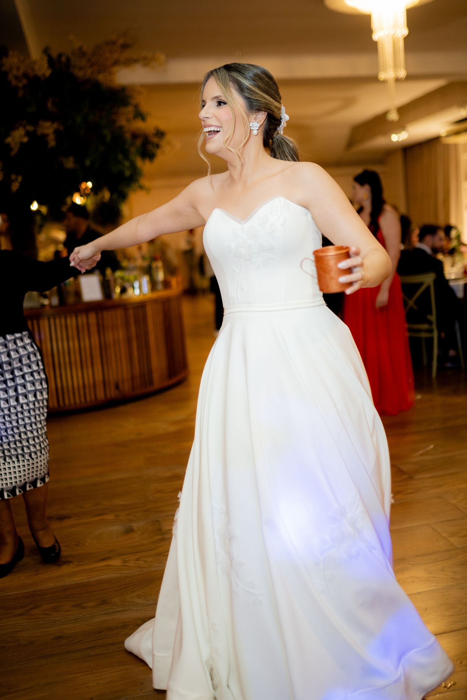 Bride in a strapless white gown dancing joyfully at her wedding reception, holding a drink and smiling.