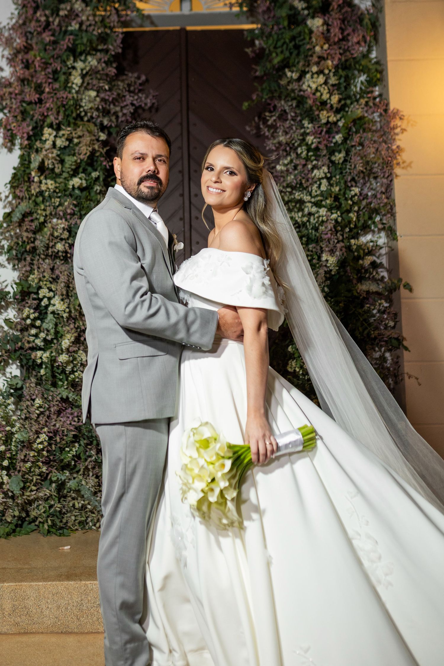 Elegant bride in white off-shoulder gown with veil holds calla lily bouquet beside groom in gray suit before floral backdrop.
