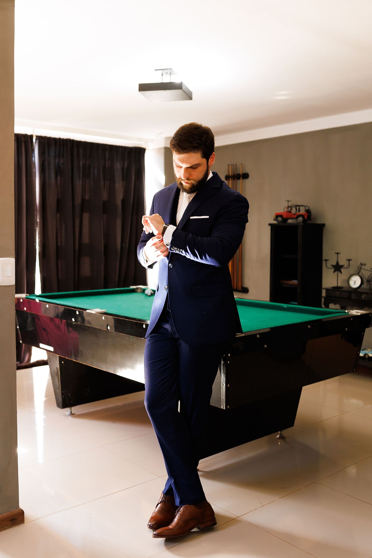 A well-dressed man in a navy blue suit adjusts his cufflinks while leaning against a green billiard table indoors.