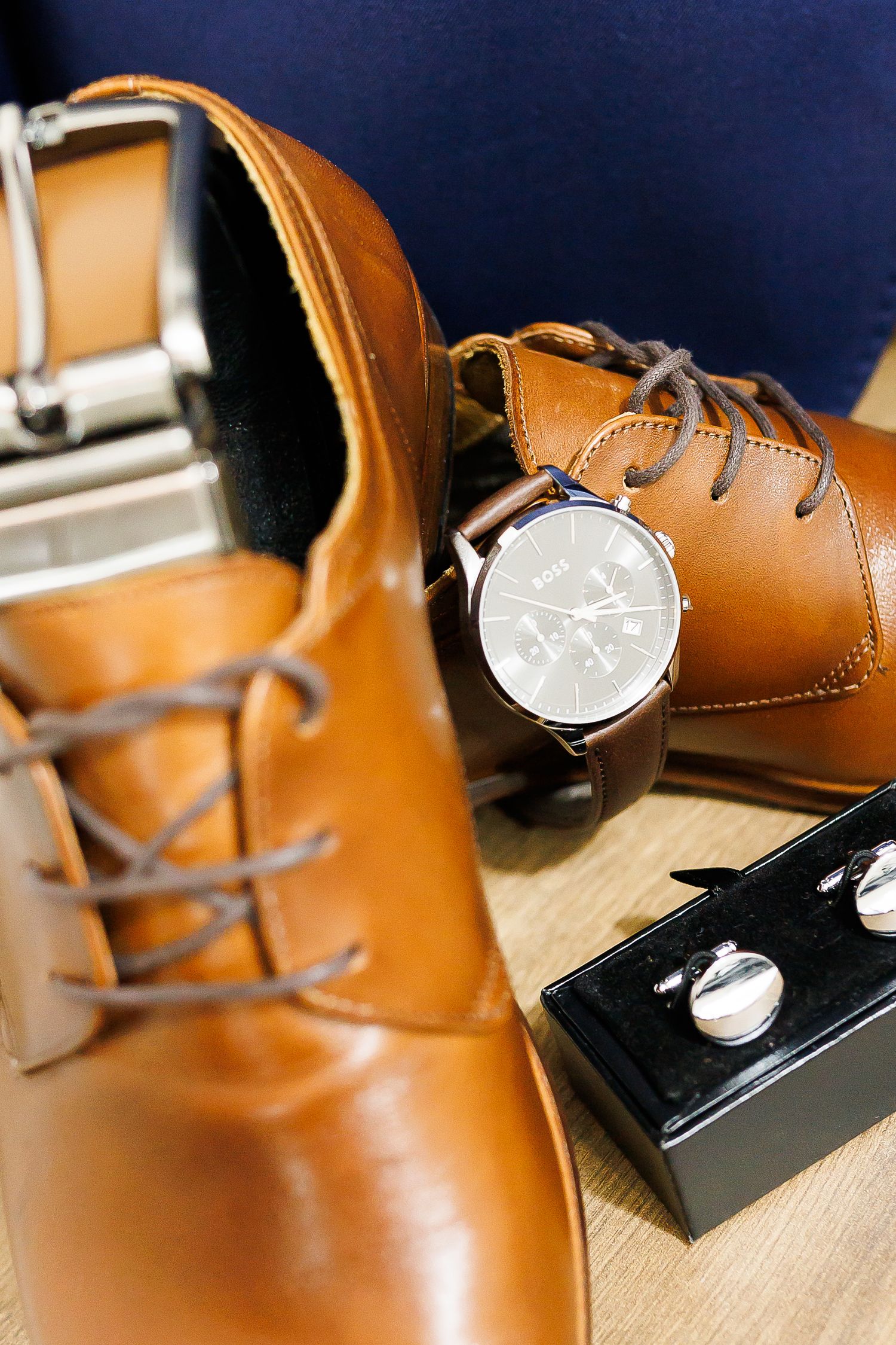 Tan leather oxford shoes with a brown leather watch and silver cufflinks on a wooden surface.