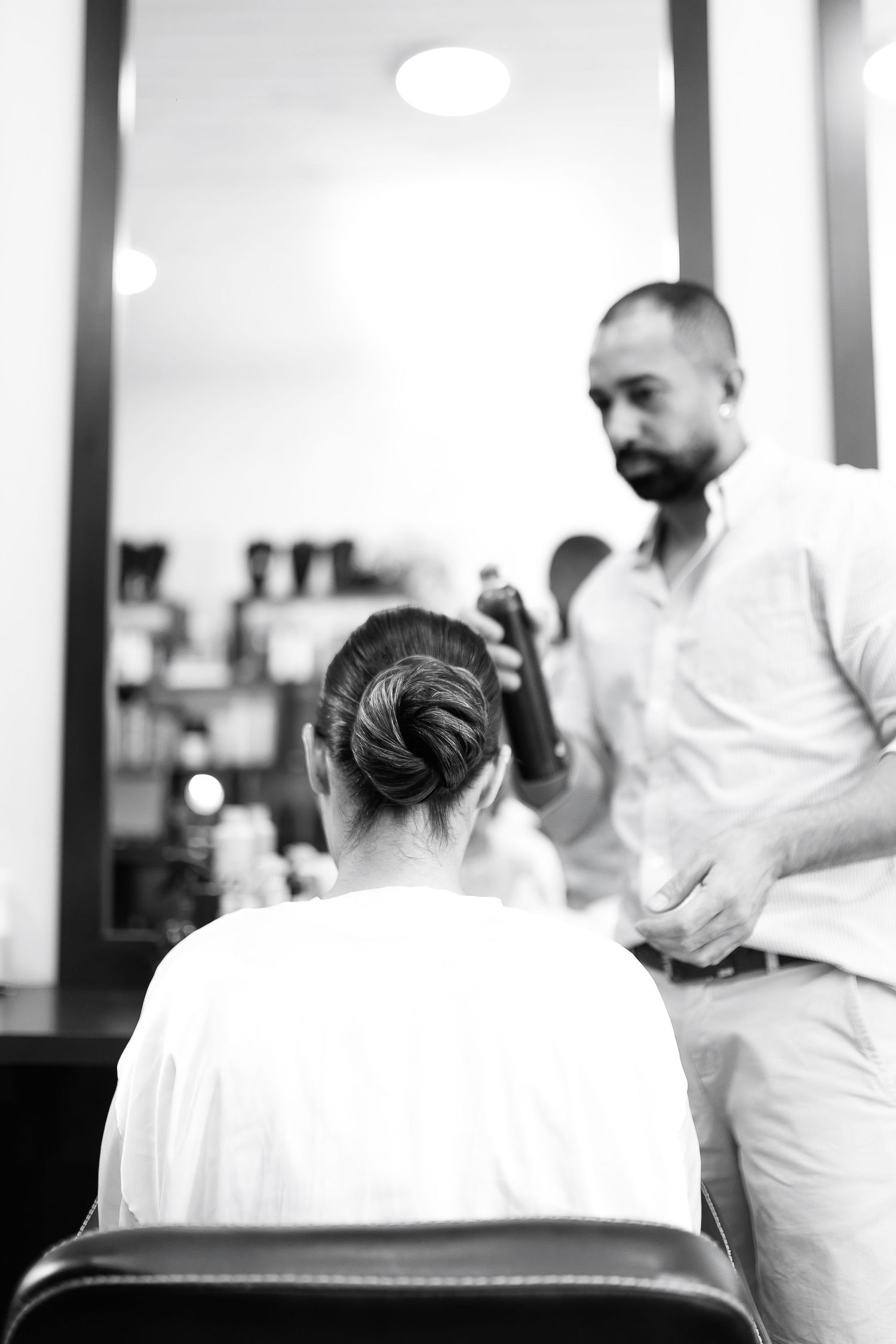 Black and white photo of a hairdresser styling a woman's hair bun in a salon chair.