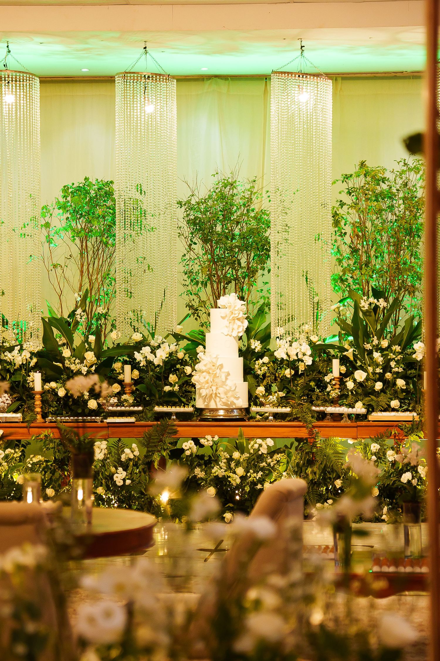 Elegant wedding reception with tiered white cake surrounded by lush greenery, white flowers, and crystal chandeliers in a warmly lit venue.