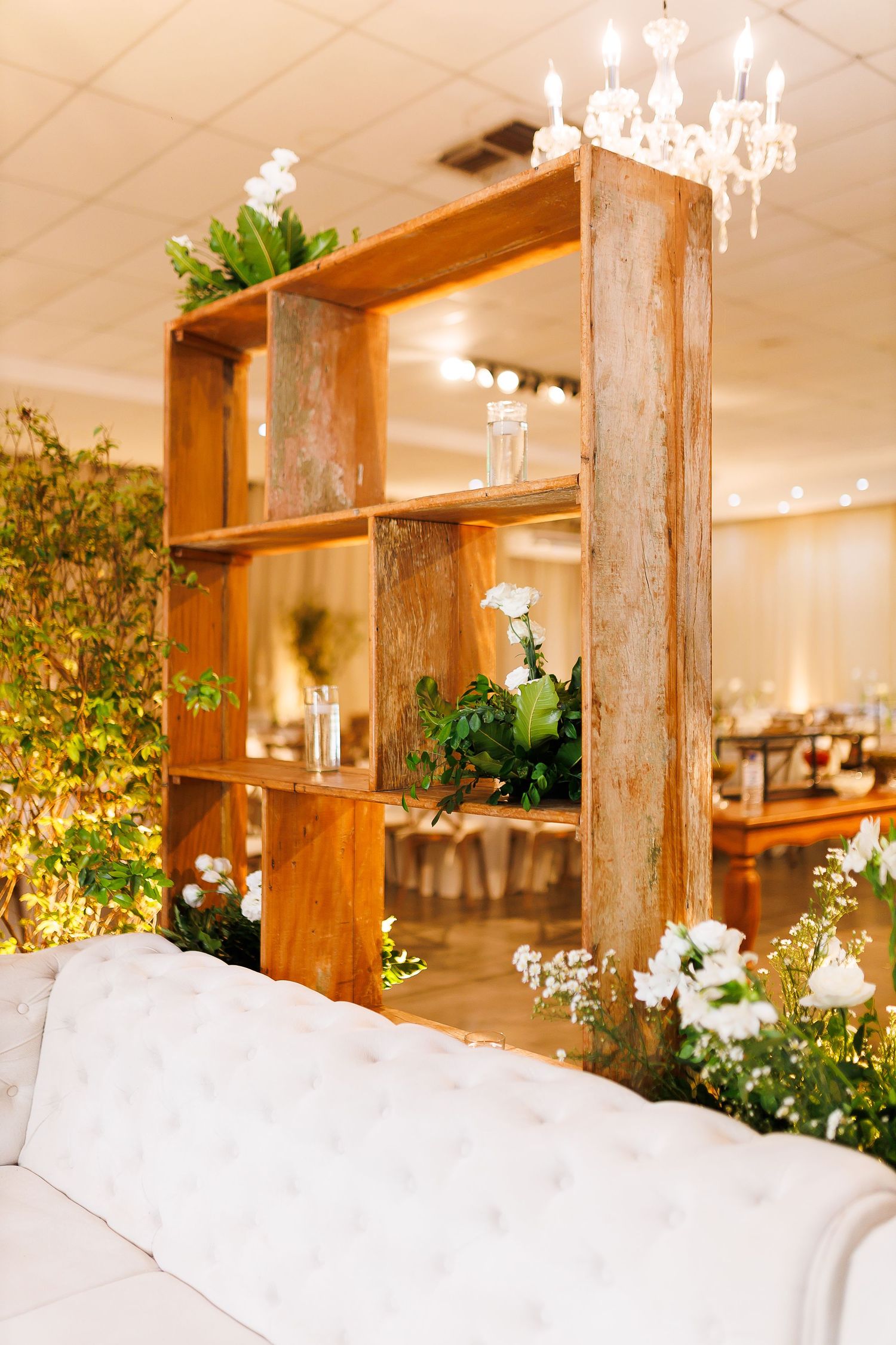 Rustic wooden shelving unit with white flowers and greenery beside a tufted white sofa in an elegant event venue.