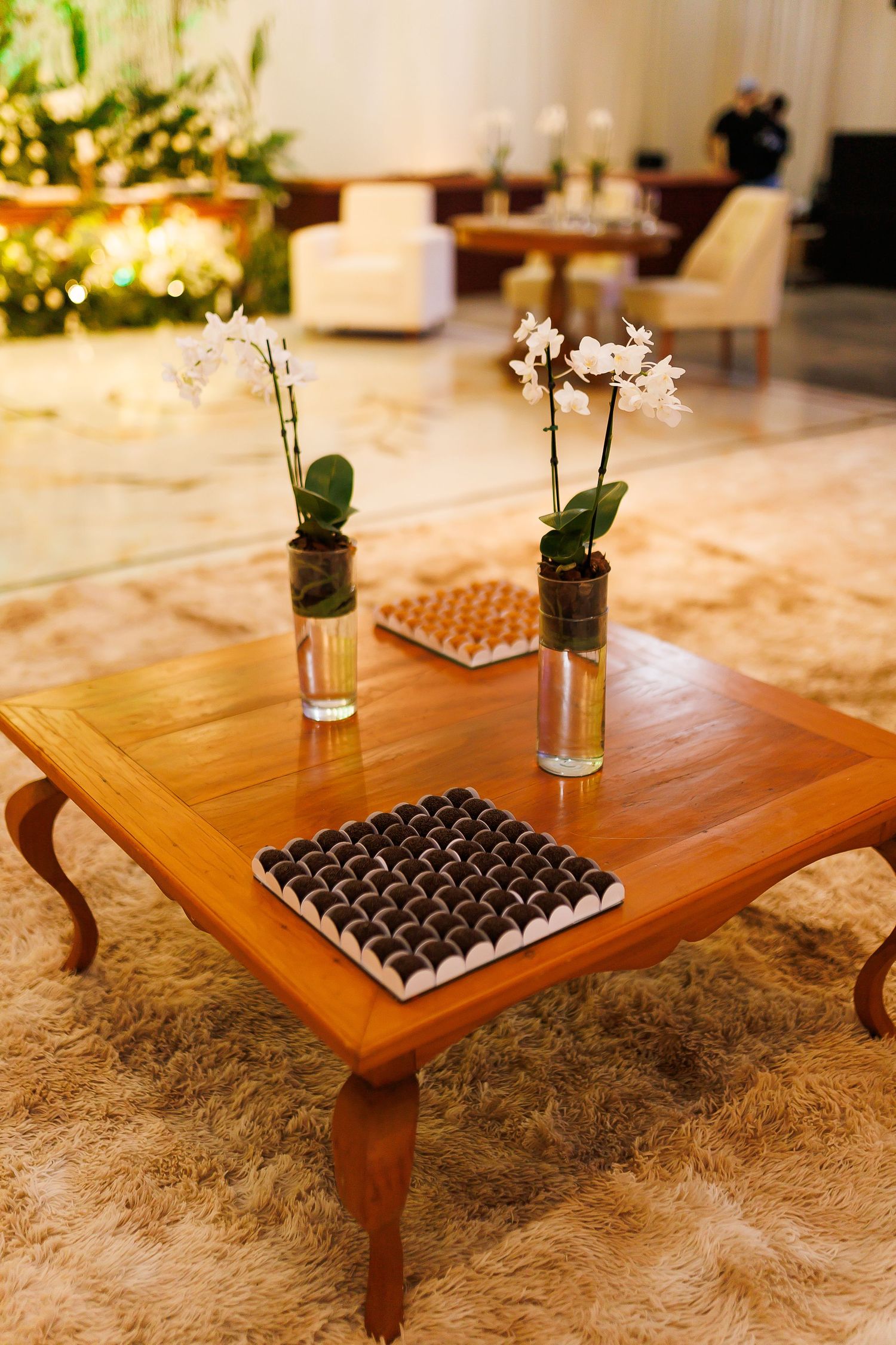 Elegant wooden coffee table with white orchids in glass vases and chocolate truffles on a plush rug.