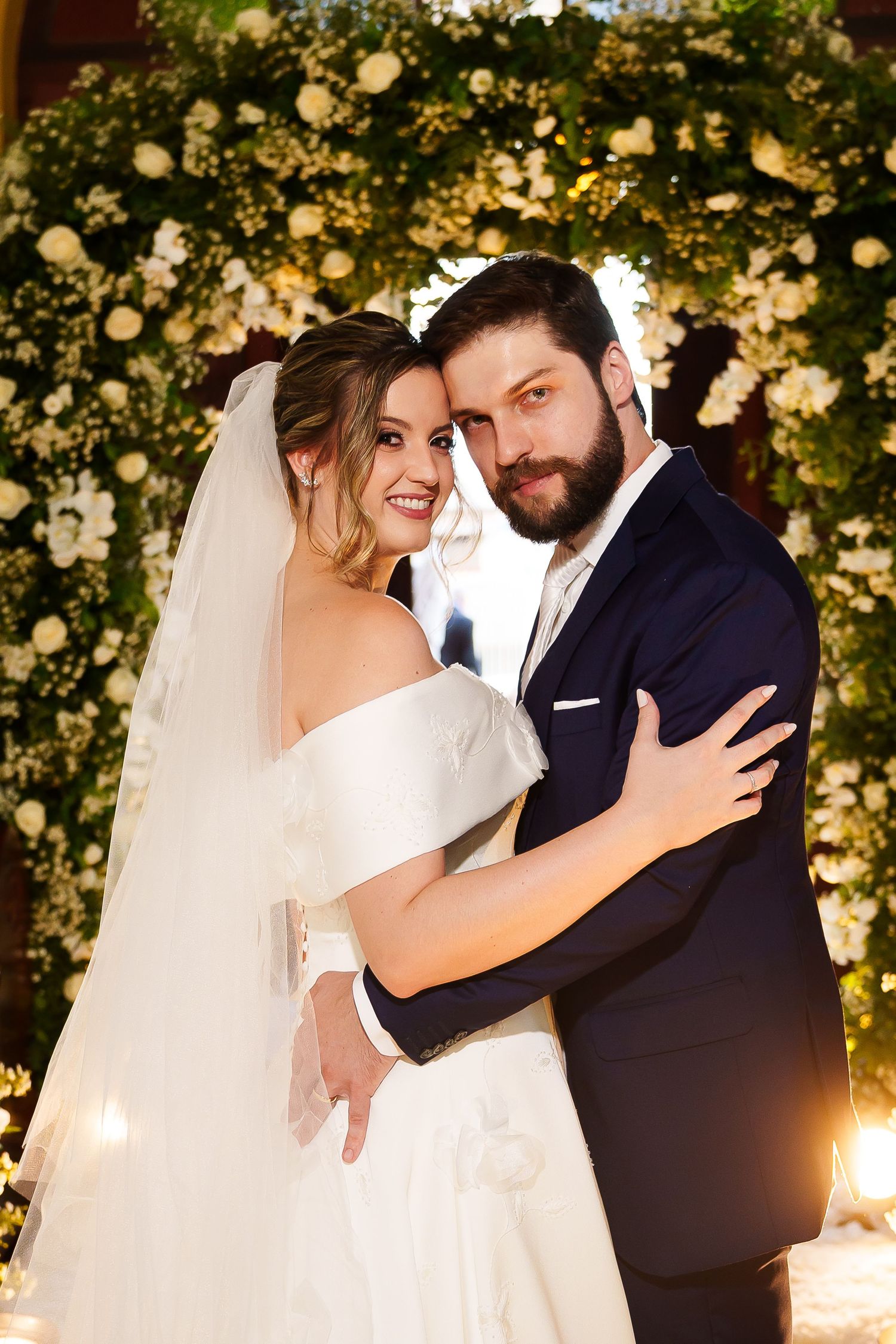 Bride in white off-shoulder gown and veil embraces bearded groom in navy suit before floral arch backdrop.