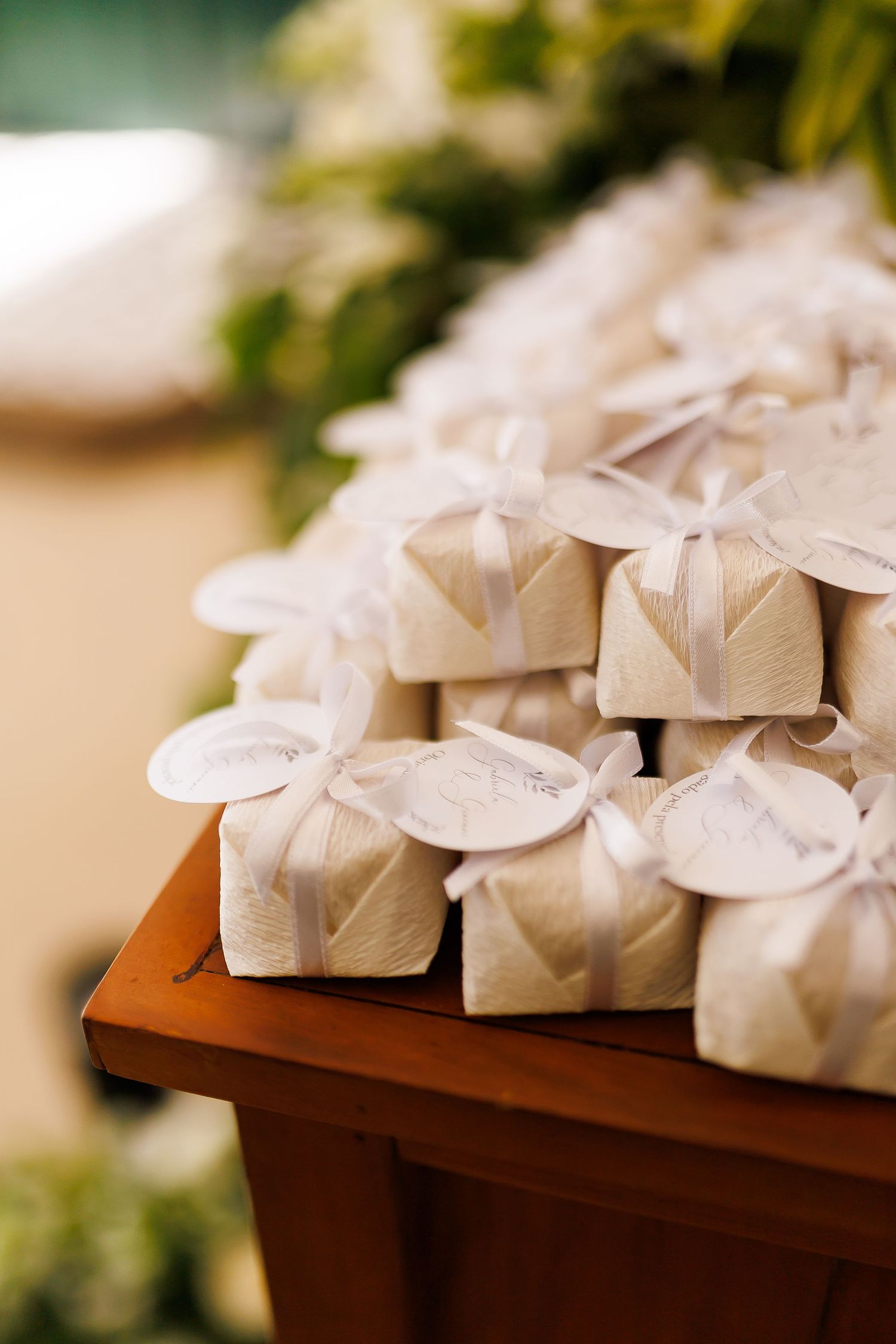 Elegant white wedding favor boxes tied with ribbon and tags, stacked on a wooden table with greenery in the background.