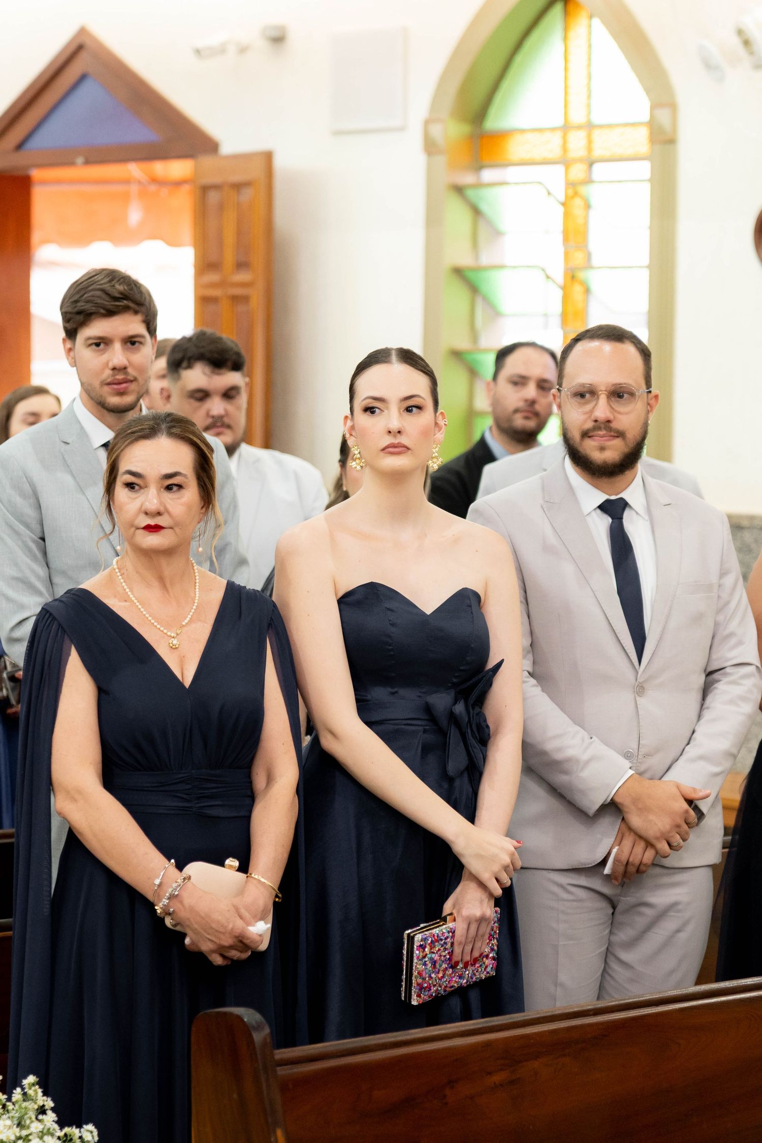 Elegant wedding guests in navy dresses and suit stand attentively in a church aisle during a ceremony.
