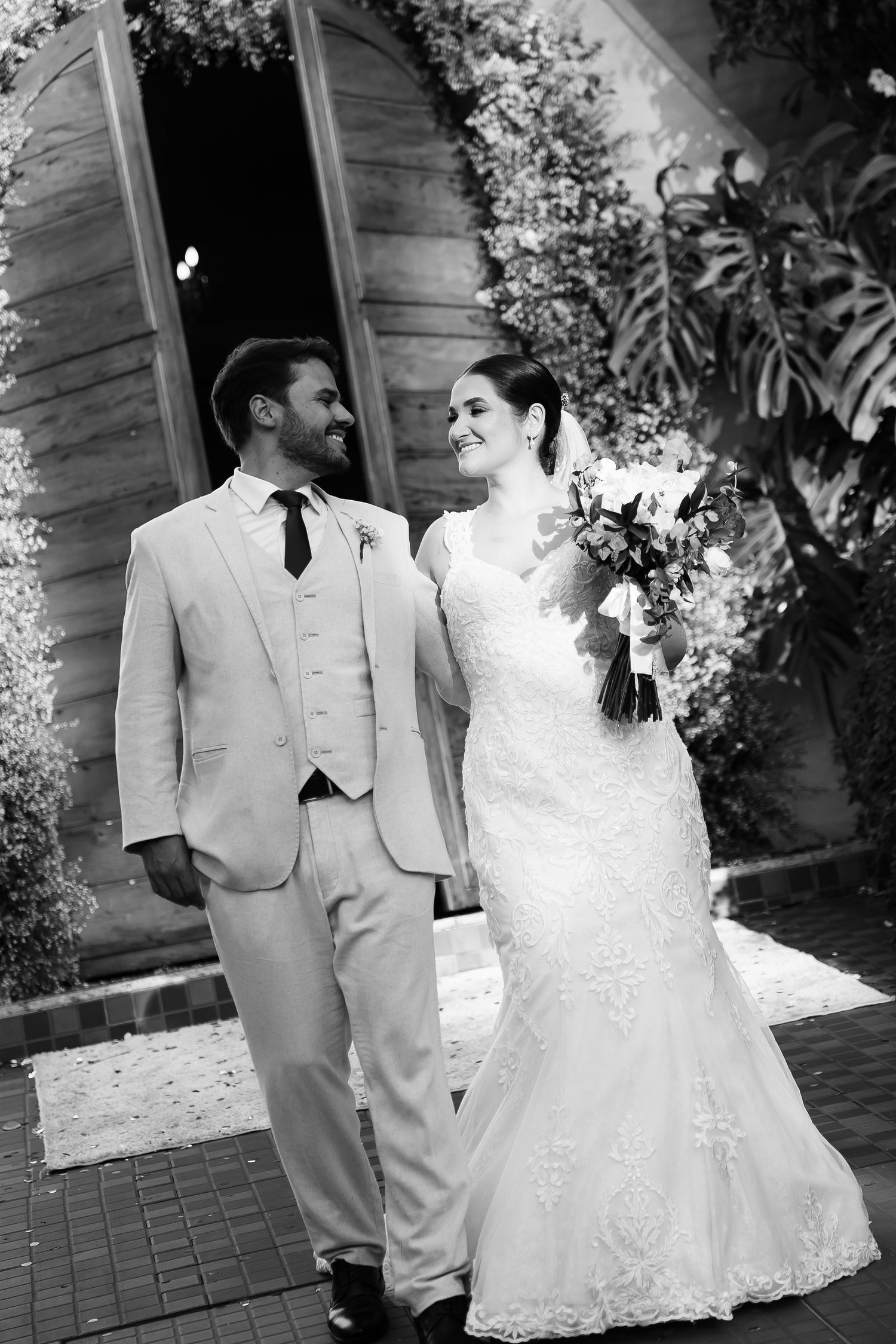 Black and white photo of a smiling bride in lace gown holding bouquet with groom in light suit on wedding day.