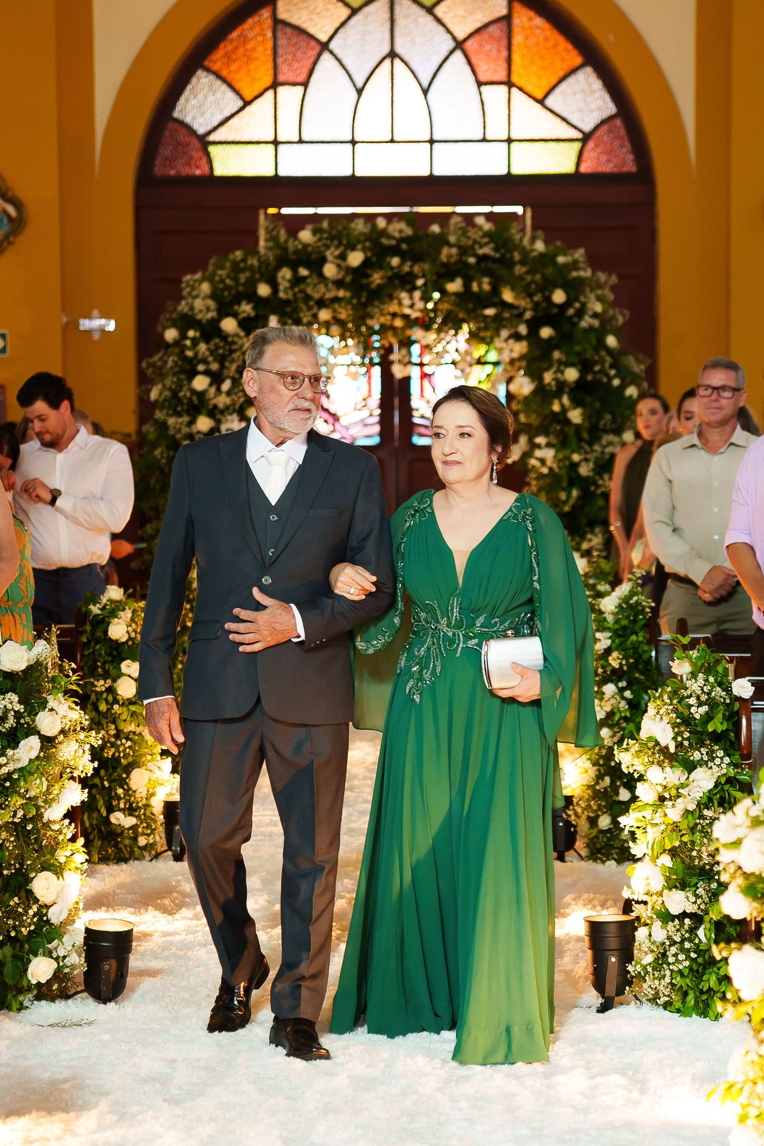 Elegant couple walking down a floral-adorned church aisle, man in gray suit, woman in emerald green gown.