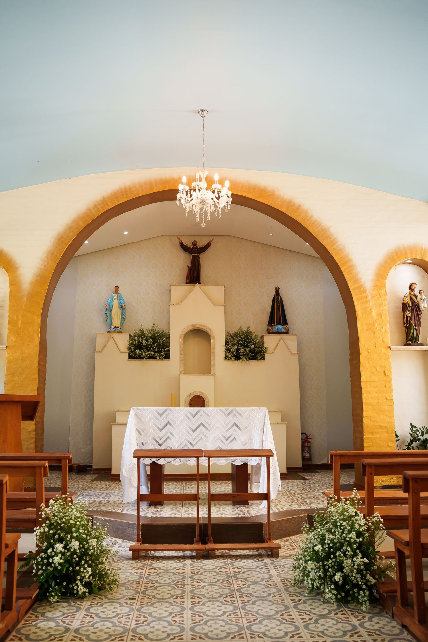 Small Catholic chapel interior with wooden pews, floral arrangements, chandelier, and altar featuring religious statues under arched alcove.