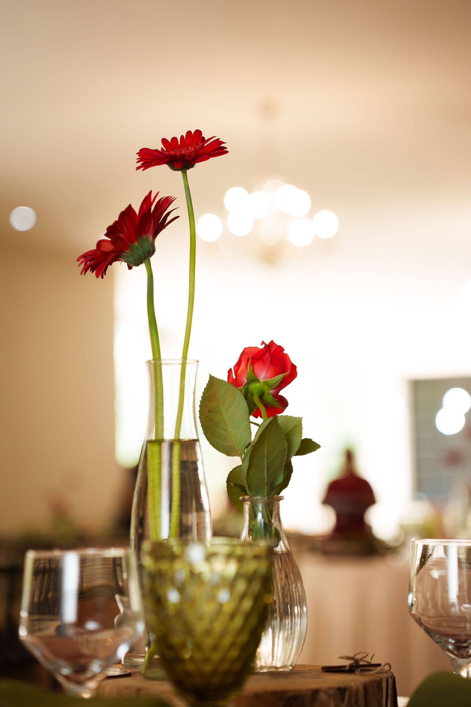 Red gerbera daisies and a rose in glass vases on an elegantly set dining table with wine glasses.