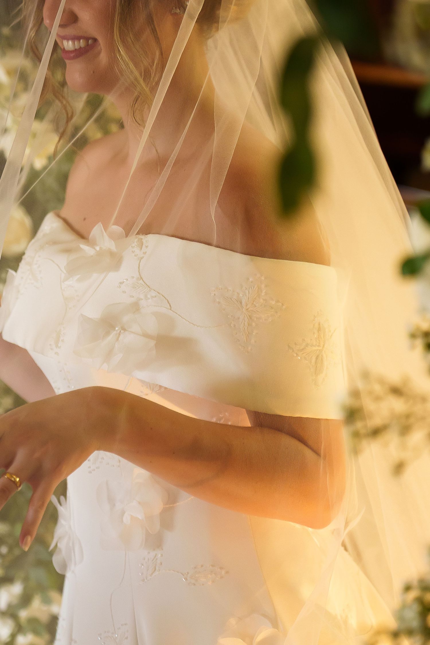 Bride in off-shoulder embroidered white gown with sheer veil, smiling in warm golden light.