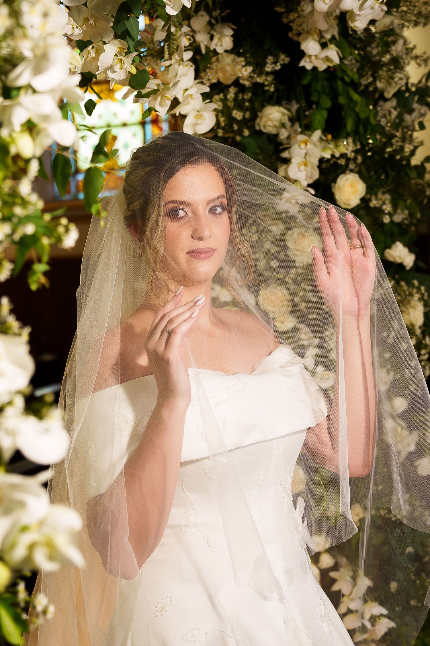 Elegant bride in white off-shoulder gown and veil poses among lush white floral arrangements in a church setting.