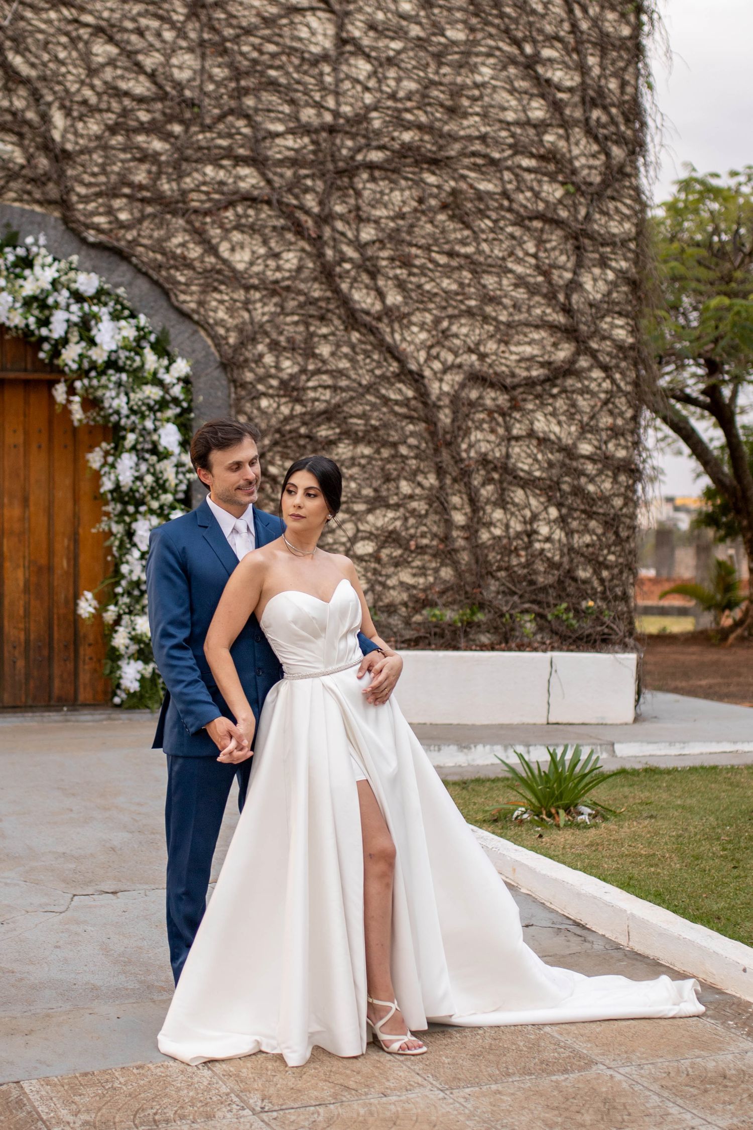 Bride in white strapless gown with slit and groom in blue suit pose together outdoors at elegant wedding venue.