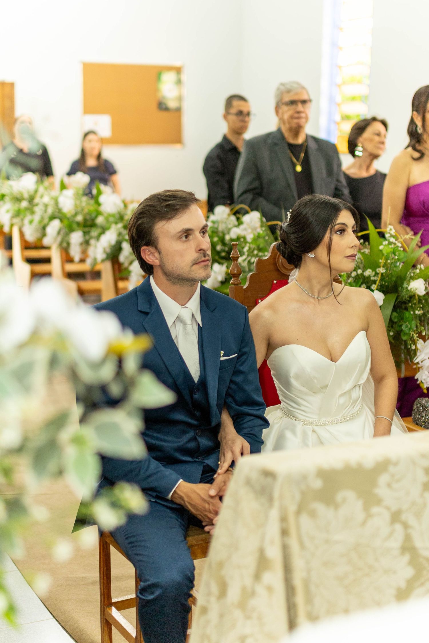 Bride in white strapless gown and groom in navy suit sit together at church altar during wedding ceremony.
