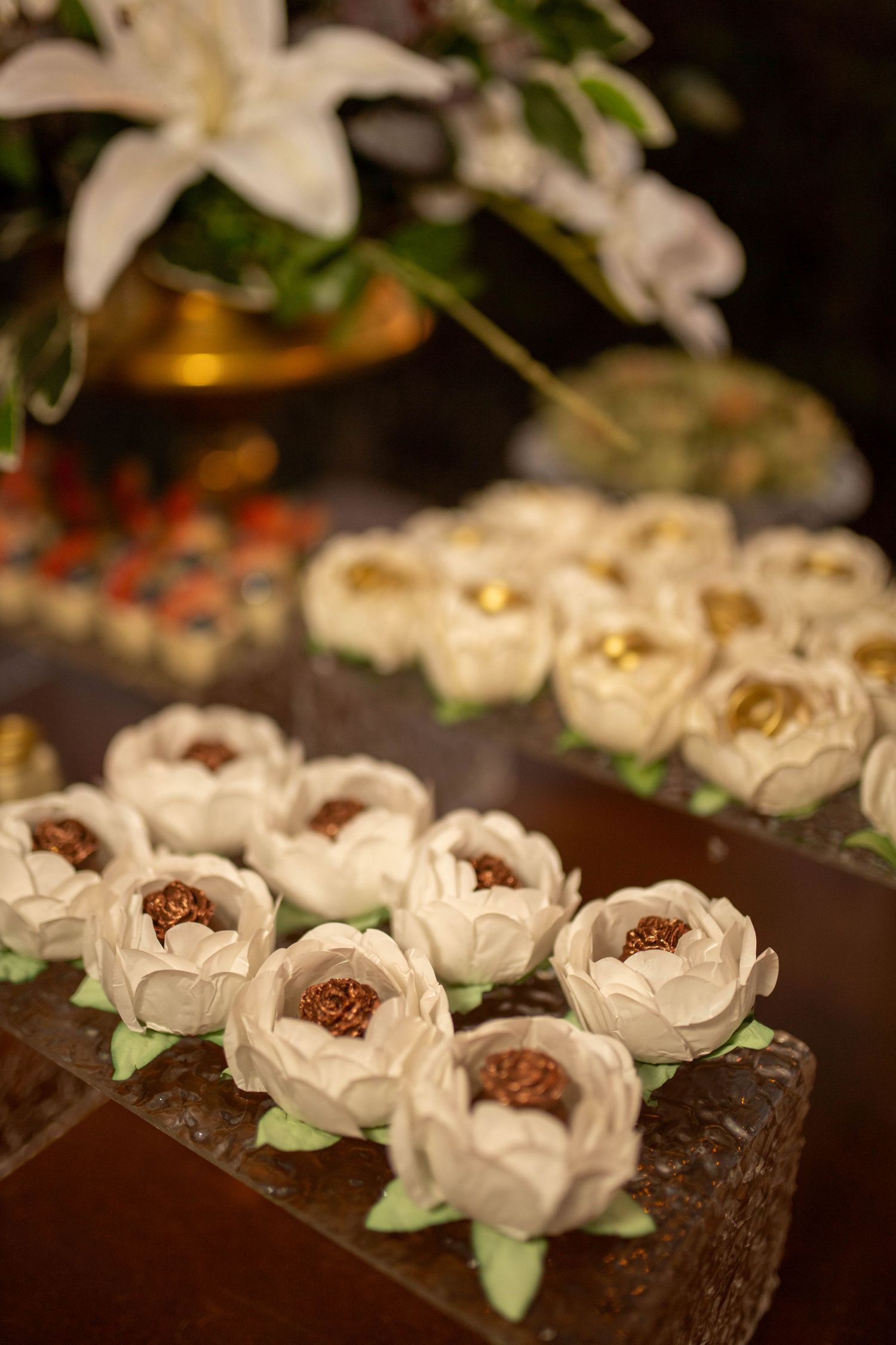 Elegant white fondant flower-shaped chocolates displayed on a dark surface with white lilies in the background.