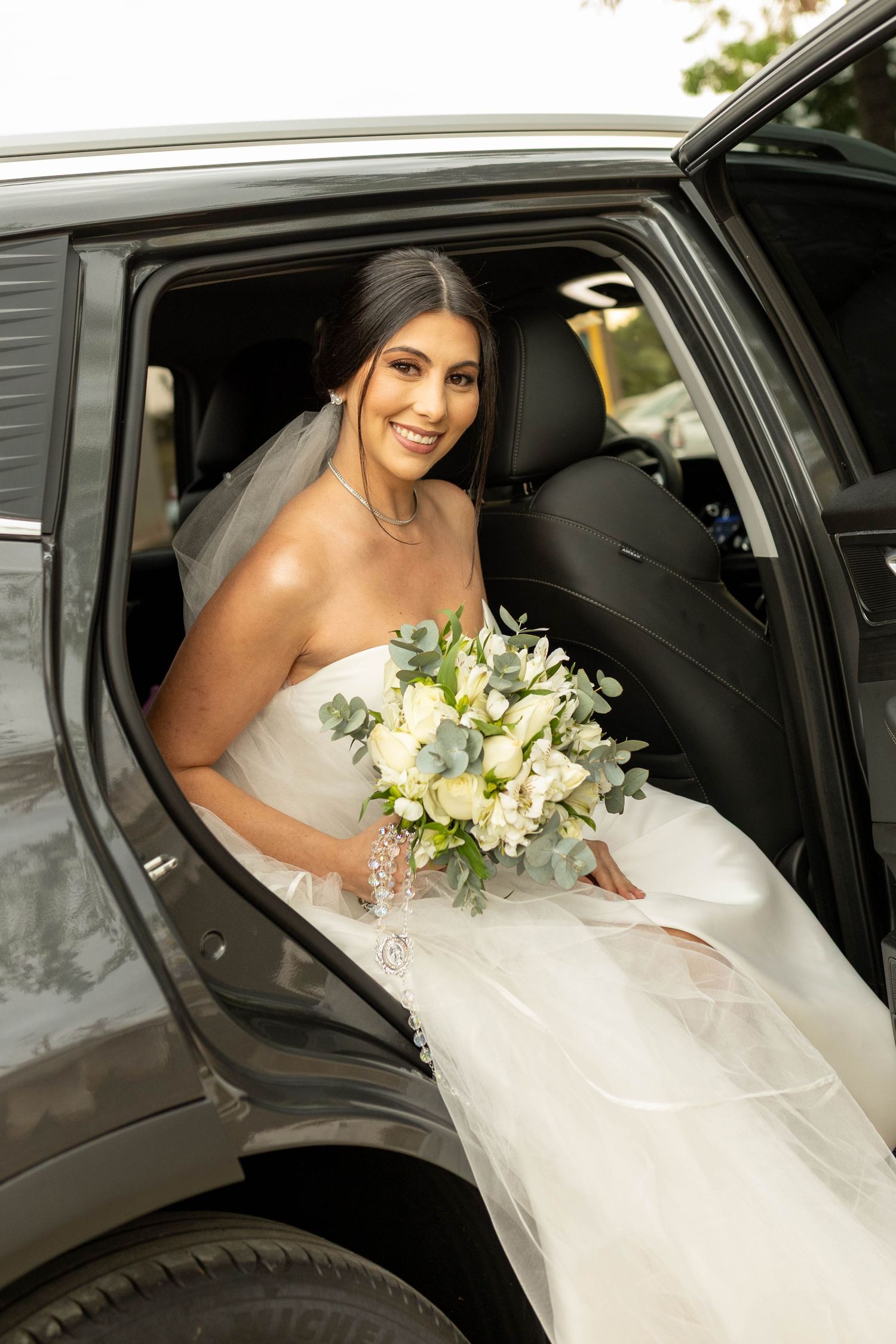 Smiling bride in strapless white gown holding bouquet of white flowers and greenery while sitting in a luxury vehicle.